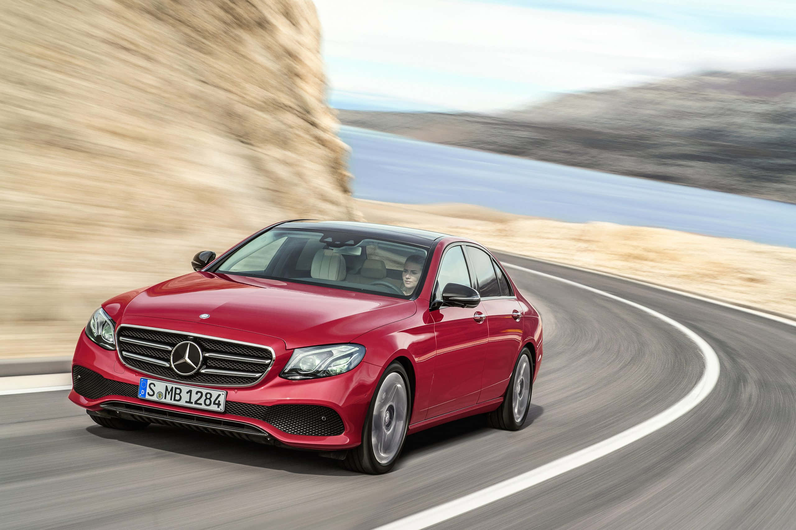 Mercedes Benz Clase E 桌布 壁紙 — 一輛鮮紅色的梅賽德斯轎車沿著蜿蜒的海岸公路飛馳，背景是模糊的懸崖和海洋。