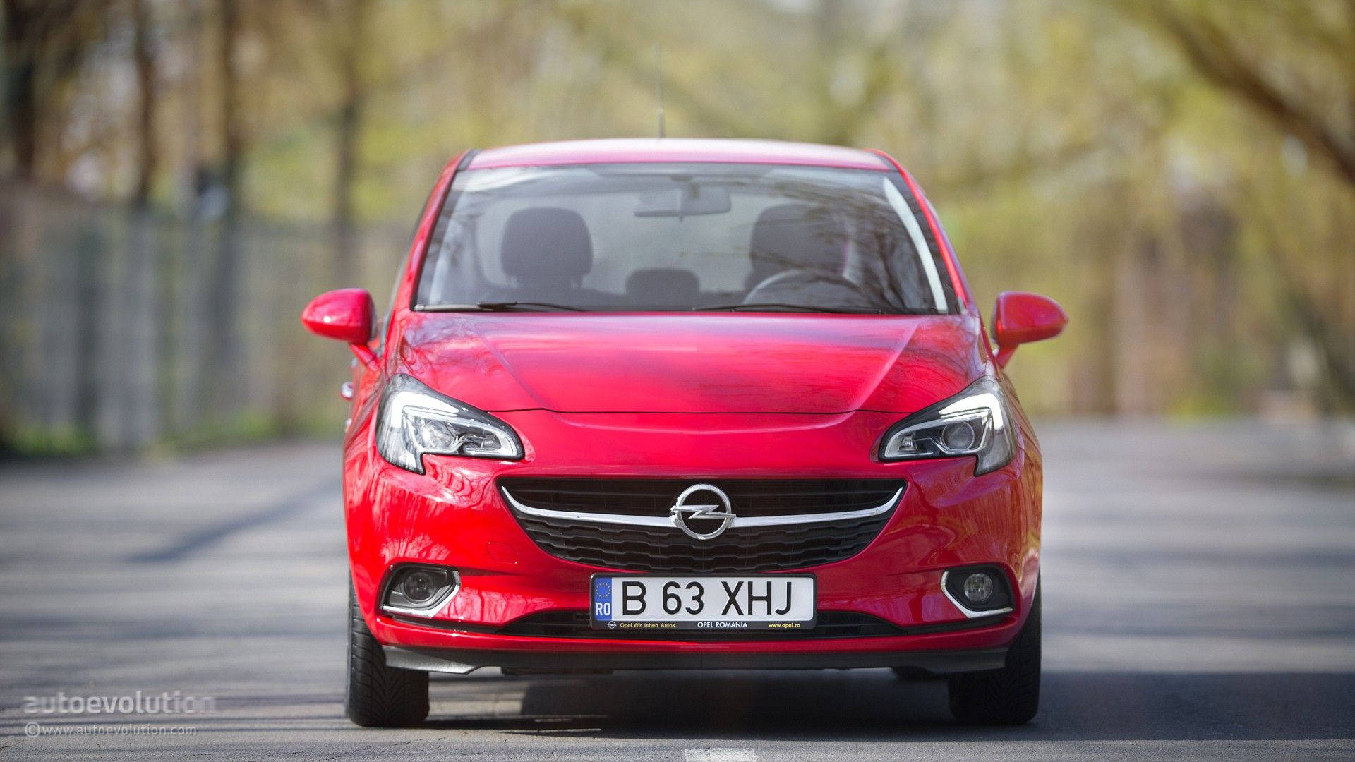 Opel 桌布 壁紙 — 一輛鮮紅色的歐寶汽車在樹木成蔭的道路上向前行駛，在明亮的日光下，背景有柔和模糊的樹葉。
