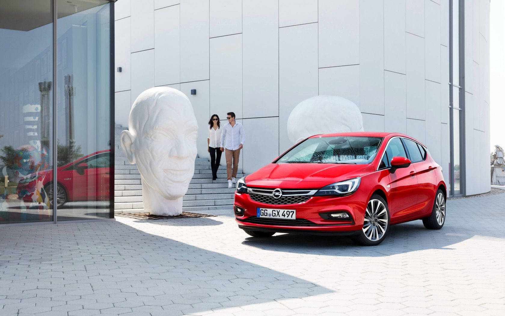 Opel 桌布 壁紙 — 一輛鮮紅色的歐寶雅特停在一棟現代建築外，建築物有巨大的白色頭部雕塑，附近有兩個人在散步。