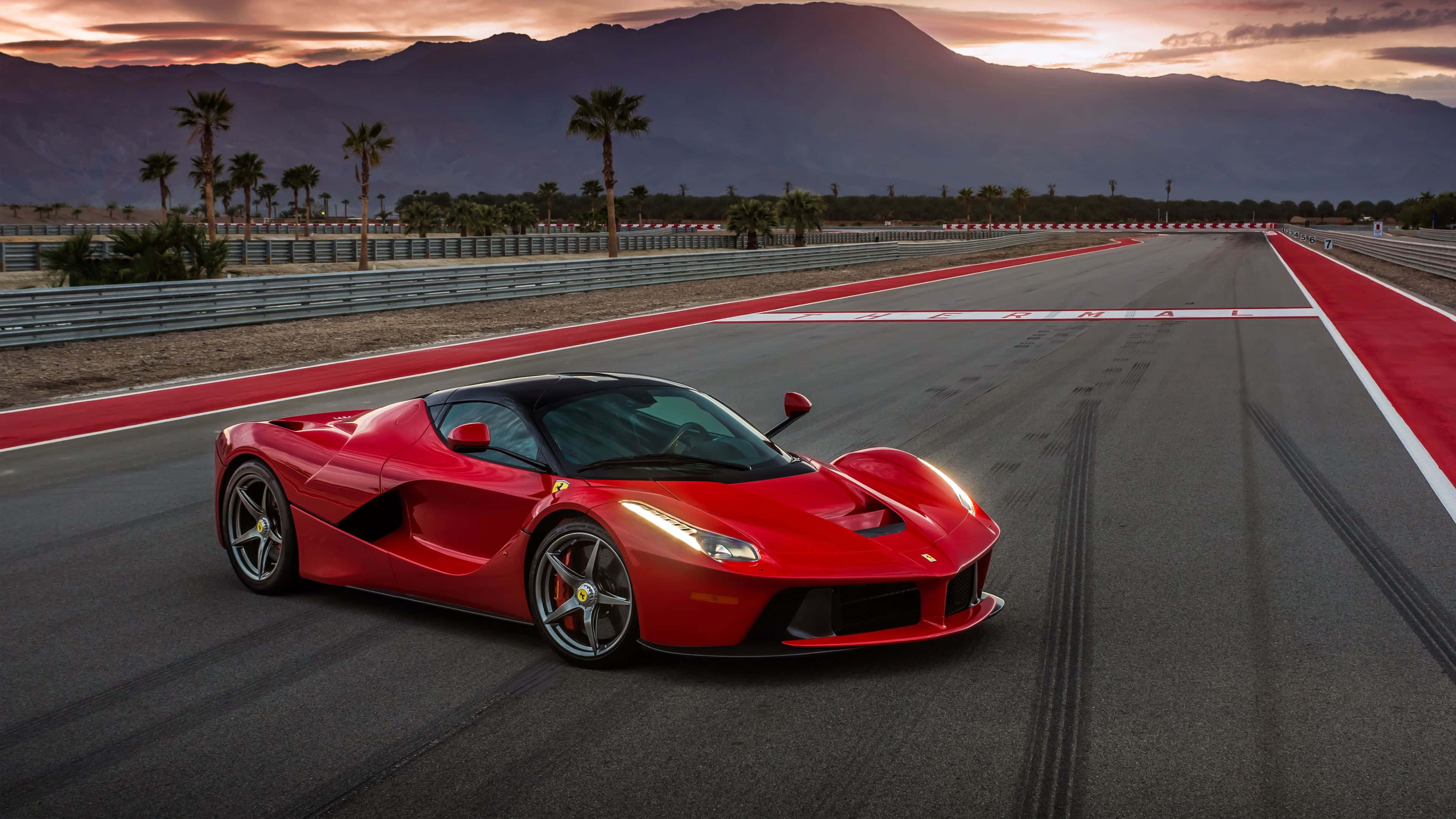 Ferrari Laferrari 桌布 壁紙 — 一輛鮮紅色的法拉利 LaFerrari 在日落時分沿著賽道飛馳，背景是山脈和棕櫚樹。