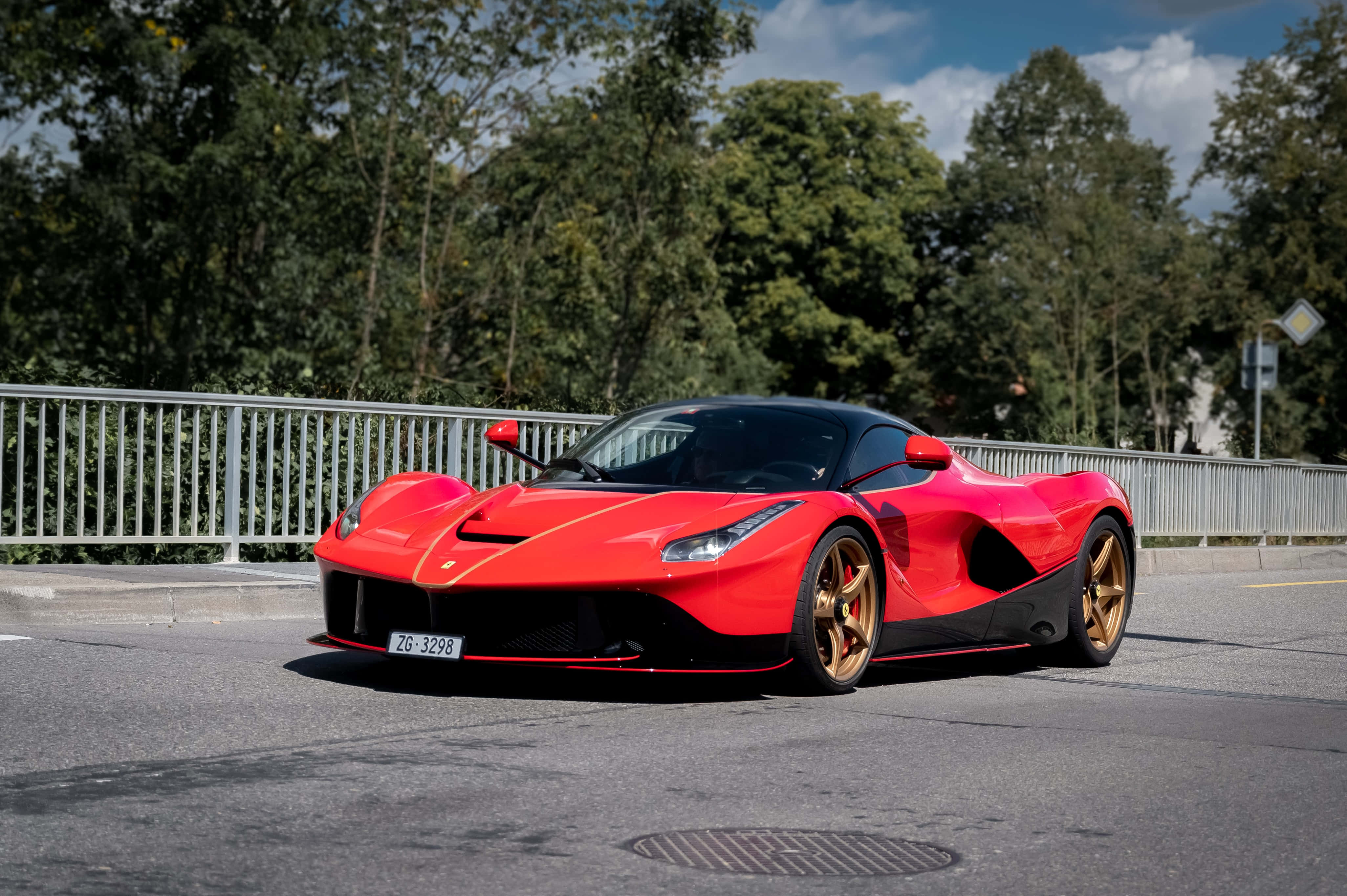 法拉利 桌布 壁紙 — 一輛鮮紅色的法拉利 LaFerrari 配金色輪圈，停泊在陽光明媚的瀝青路上，旁邊有金屬欄杆和茂密的綠樹。