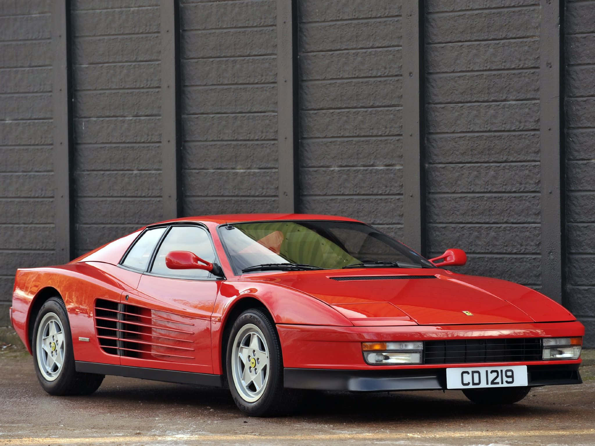 Ferrari Testarossa 桌布 壁紙 — 一輛鮮紅色的法拉利 Testarossa 在日光下閃耀，停放在有紋理的灰色工業牆前。