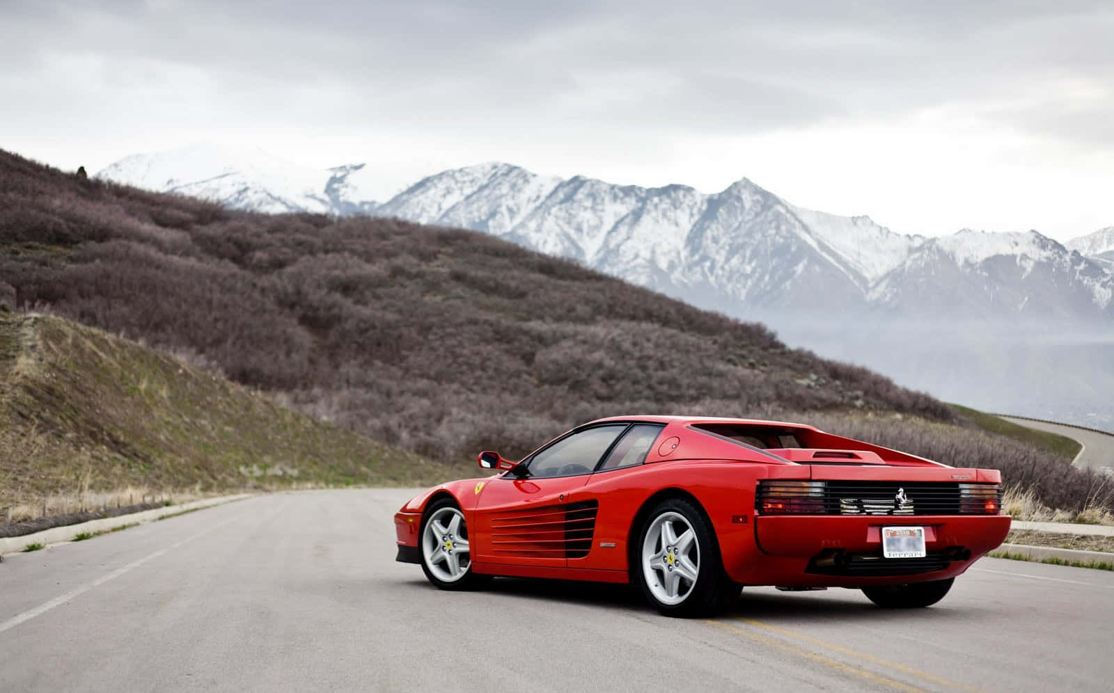Ferrari Testarossa 桌布 壁紙 — 一輛鮮紅色的法拉利 Testarossa 沿著蜿蜒的山路行駛，遠處陰雲密佈，白雪覆蓋的山峰若隱若現。