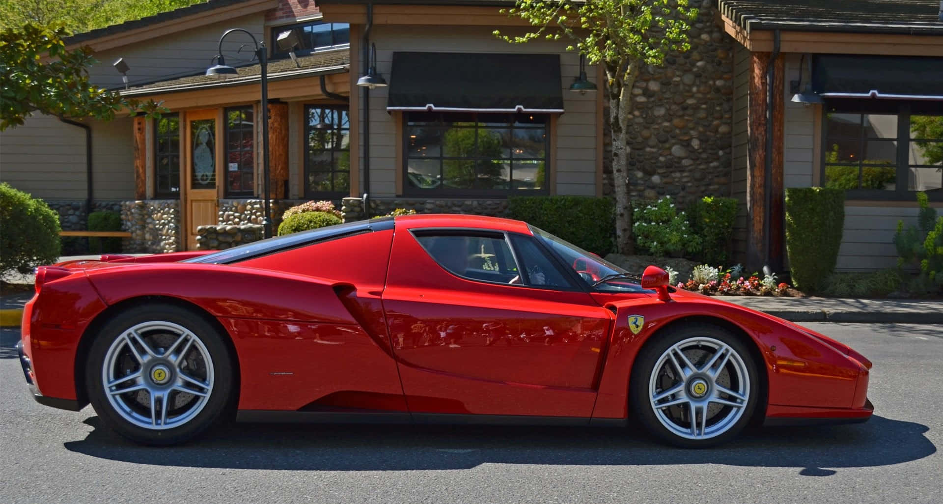 Ferrari Enzo 桌布 壁紙 — 一輛鮮紅色的法拉利Enzo停放在陽光明媚的街道上，旁邊是一座帶有綠植的質樸石木建築。