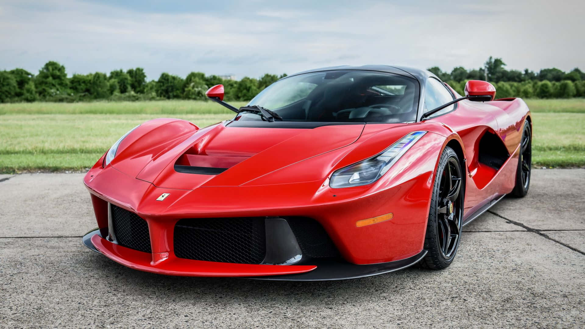 Ferrari Laferrari 桌布 壁紙 — 一輛鮮紅色的法拉利LaFerrari超級跑車在日光下，於混凝土平台上閃閃發光，背景是綠色的田野和樹木。