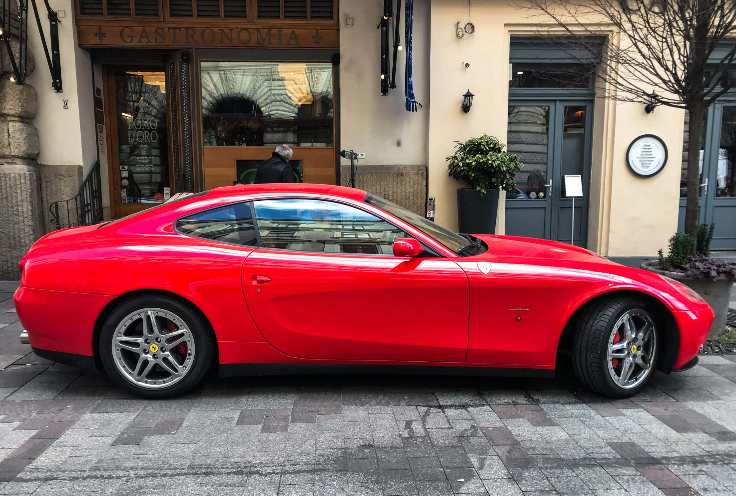 Ferrari 612 Scaglietti 桌布 壁紙 — 一輛鮮紅色的法拉利跑車停放在濕漉漉的鵝卵石街道上，前方是餐廳，周圍有光禿的樹木和柔和的白日光線。