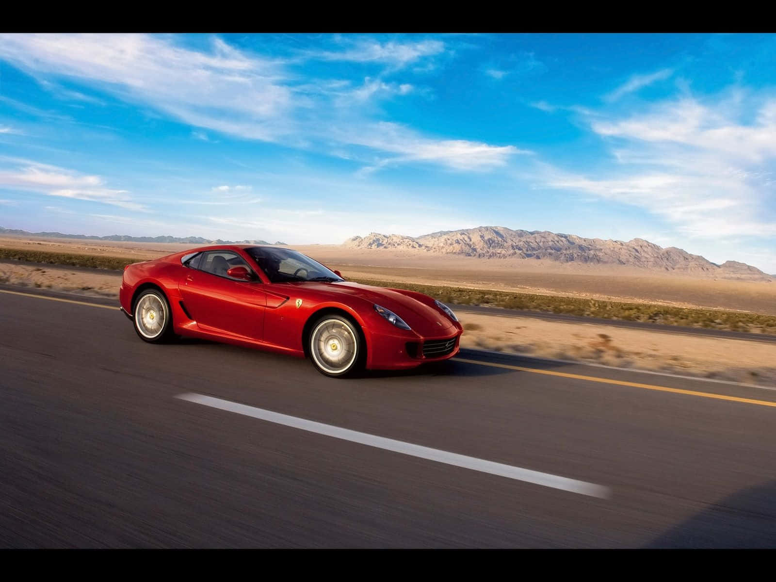 Ferrari 599 Gtb Fiorano 桌布 壁紙 — 一輛鮮紅色的法拉利跑車在明亮的藍天下，沿著沙漠高速公路疾馳，遠處有山脈。