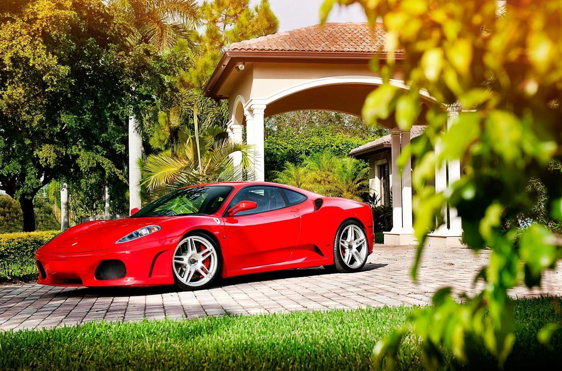 Ferrari F430 桌布 壁紙 — 一輛鮮紅色的法拉利跑車在明亮的陽光下，在鋪砌的車道上閃閃發光，車道兩旁是茂密的綠植和一座白色柱子的涼亭。