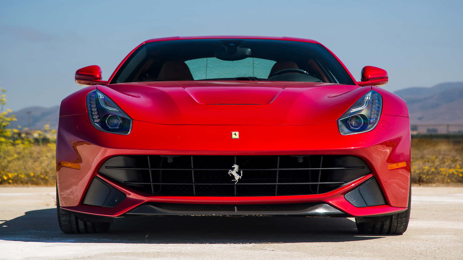 Ferrari F12 Berlinetta 桌布 壁紙 — 一輛鮮紅色的法拉利跑車在陽光明媚的道路上向前行駛，遠方有山丘，天空清澈湛藍。
