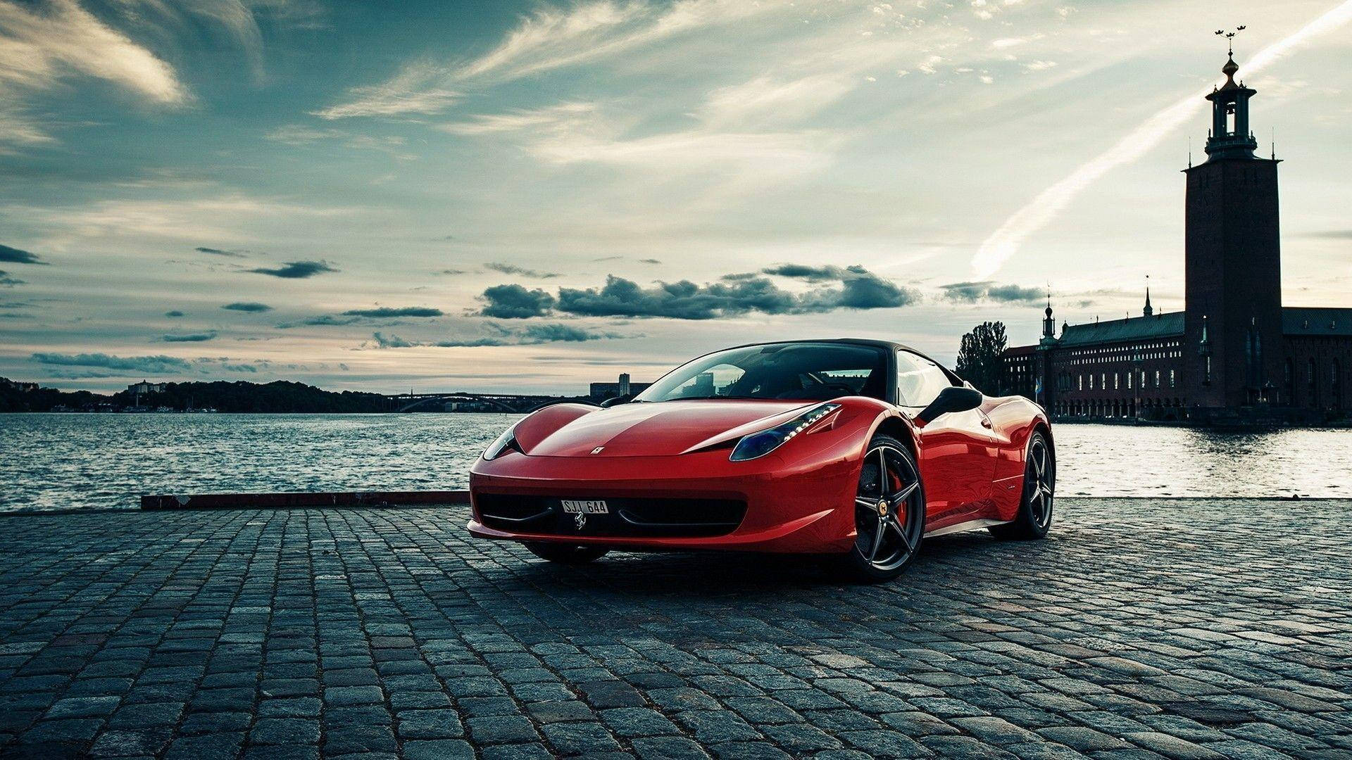 1920x1080 Ferrari 桌布 壁紙 — 一輛鮮紅色的法拉利跑車在黃昏時分於鵝卵石海濱閃閃發光，背景是歷史悠久的塔樓和寧靜的水面。