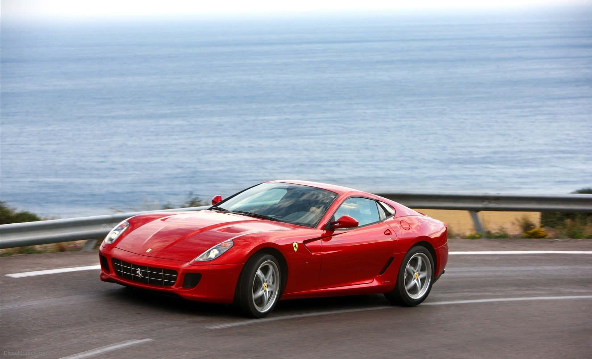 Ferrari 599 Gtb Fiorano 桌布 壁紙 — 一輛鮮紅色的法拉利跑車沿著海岸道路行駛，背後是廣闊的藍色海洋。