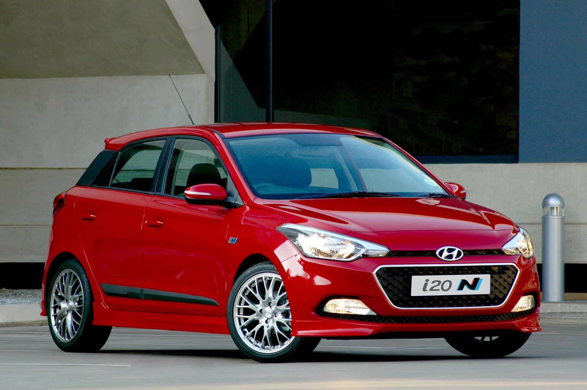 Hyundai 桌布 壁紙 — 一輛鮮紅色的現代 i20 N 掀背車停在現代建築前，配備時尚合金輪圈和運動化設計。