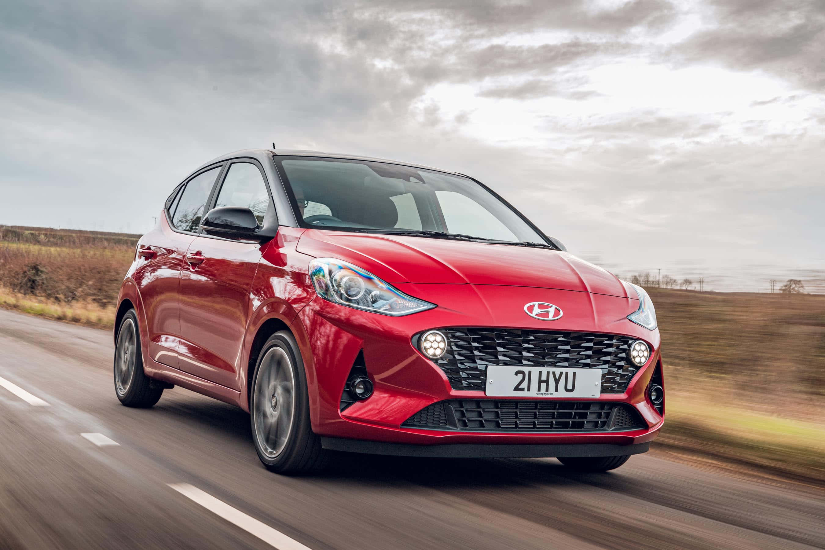 Hyundai 桌布 壁紙 — 一輛鮮紅色的現代 i20 在陰天下的鄉村道路上飛馳，背景呈現動態模糊效果。