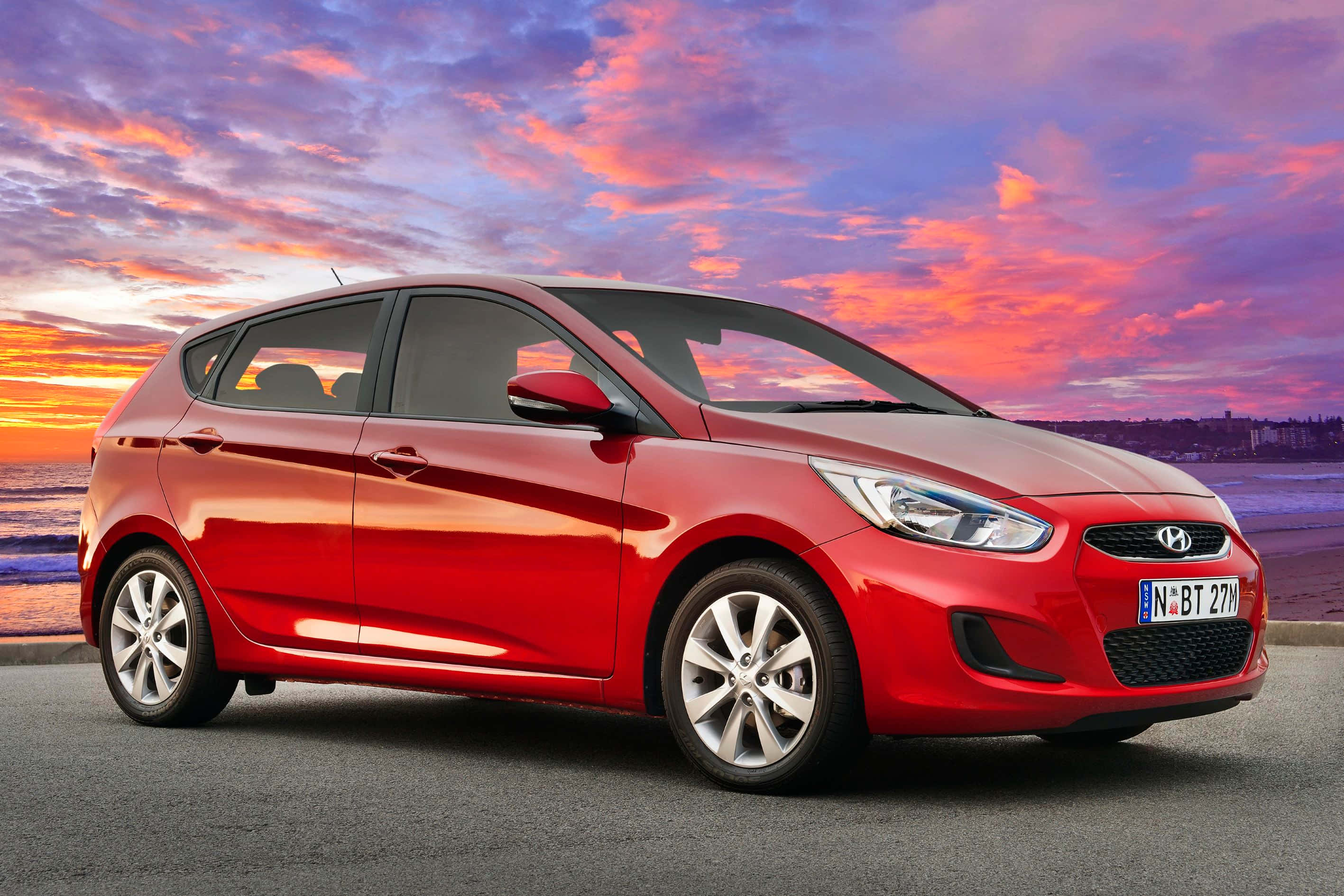 Hyundai 桌布 壁紙 — 一輛鮮紅色的現代 i20 掀背車在橙、粉紅和紫色雲彩的熾熱日落天空下，沿海岸道路閃閃發光。