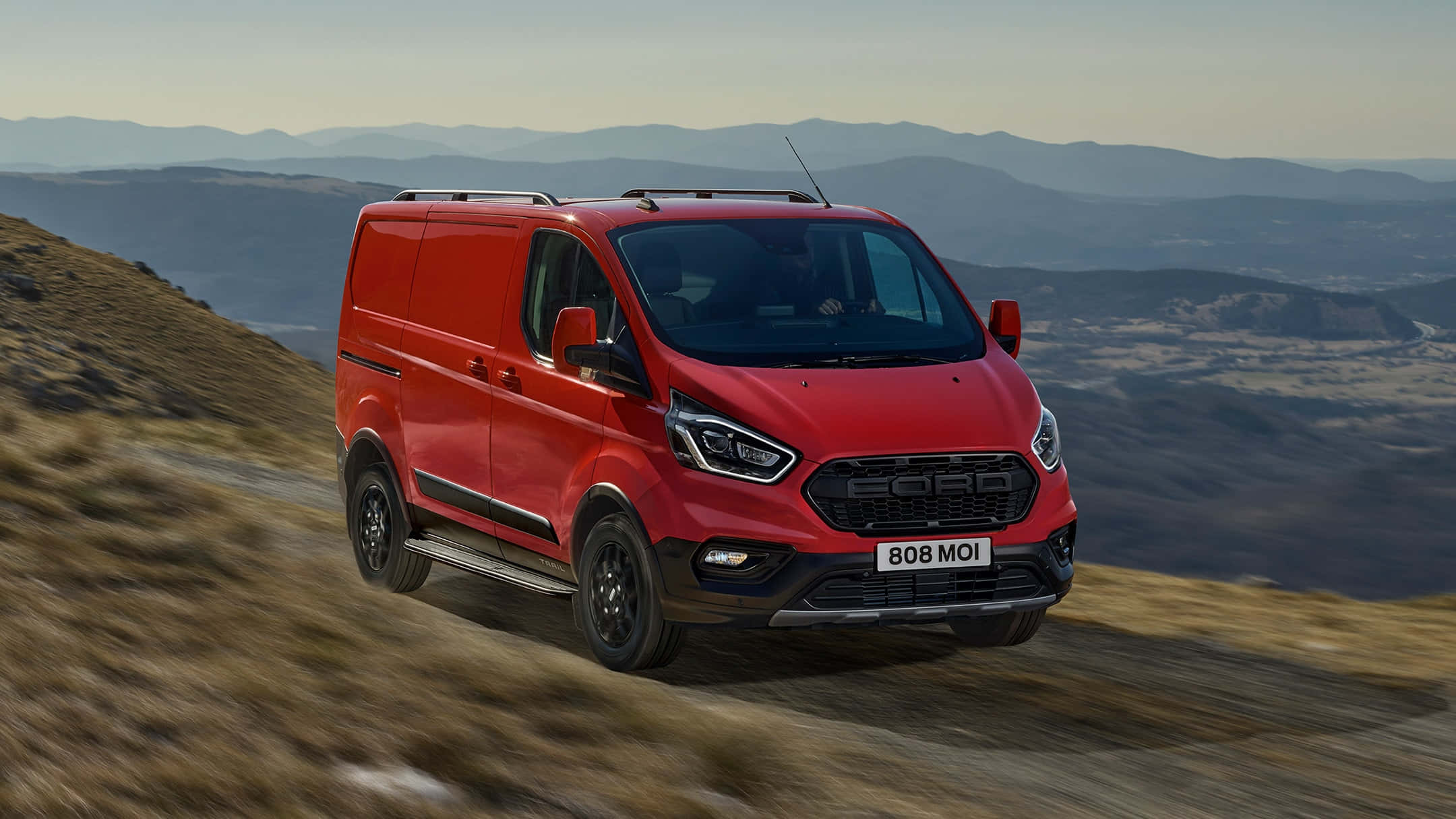 Ford Transit 桌布 壁紙 — 一輛鮮紅色的福特全順廂型車在崎嶇的山路上行駛，背景是模糊的地形和遠處的山峰，天空朦朧。