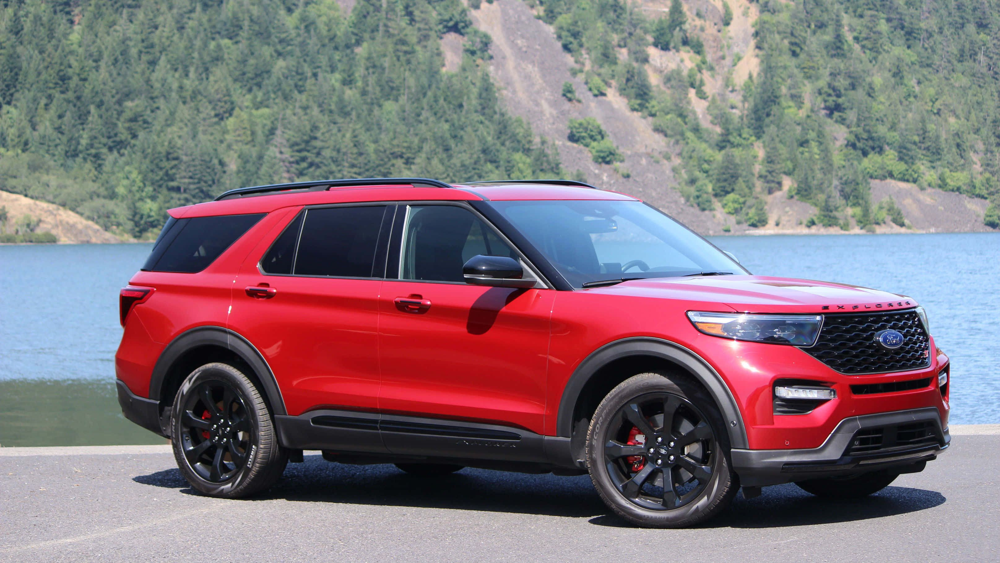 Ford Explorer 桌布 壁紙 — 一輛鮮紅色的福特探險者SUV，帶有黑色飾邊，停在一個寧靜的湖邊，周圍是森林覆蓋的山丘，在明亮的日光下。