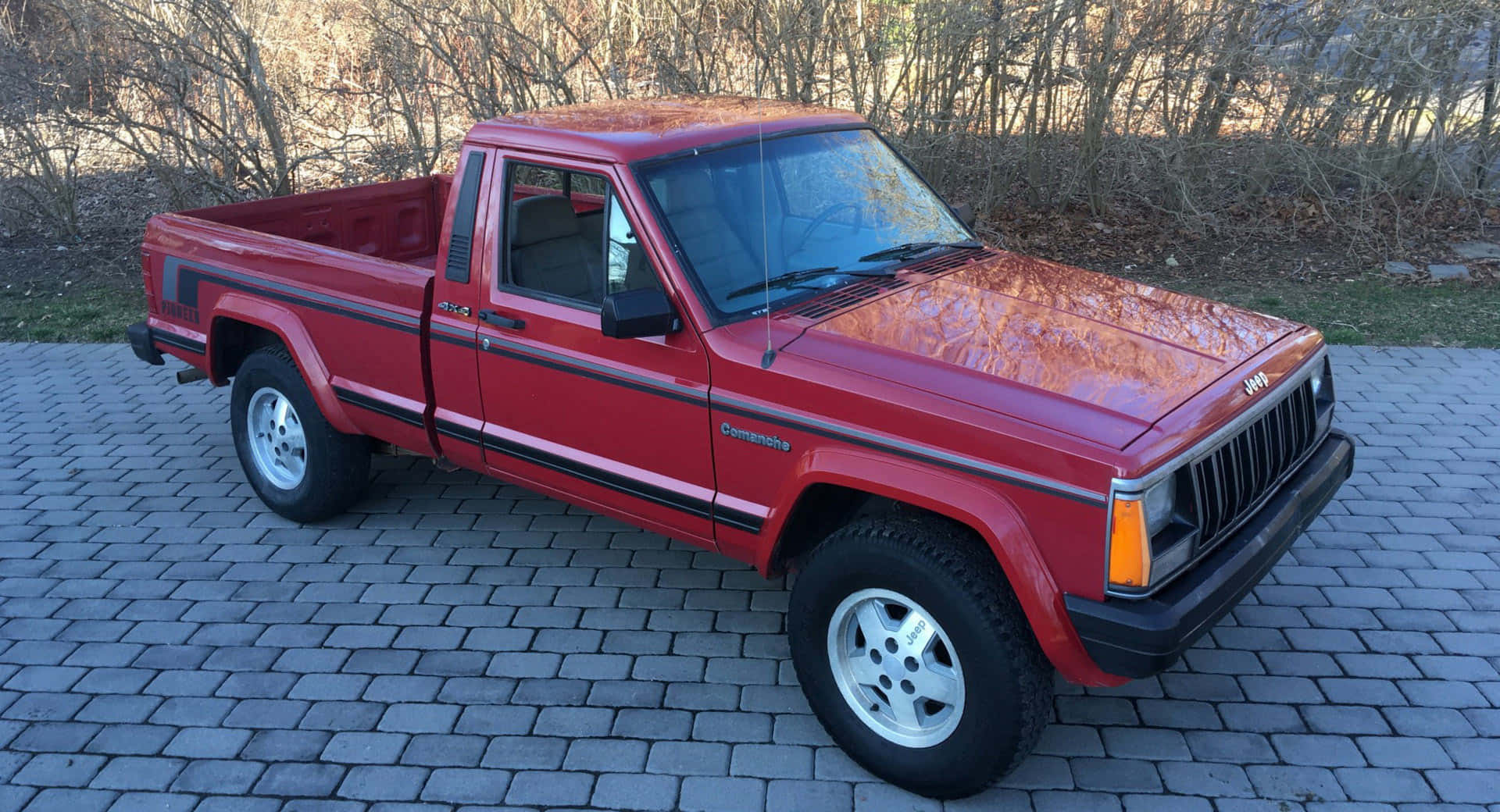 Jeep Comanche 桌布 壁紙 — 一輛鮮紅色的經典Jeep Comanche皮卡車停在灰色磚砌車道上，背景是光禿禿的樹木。