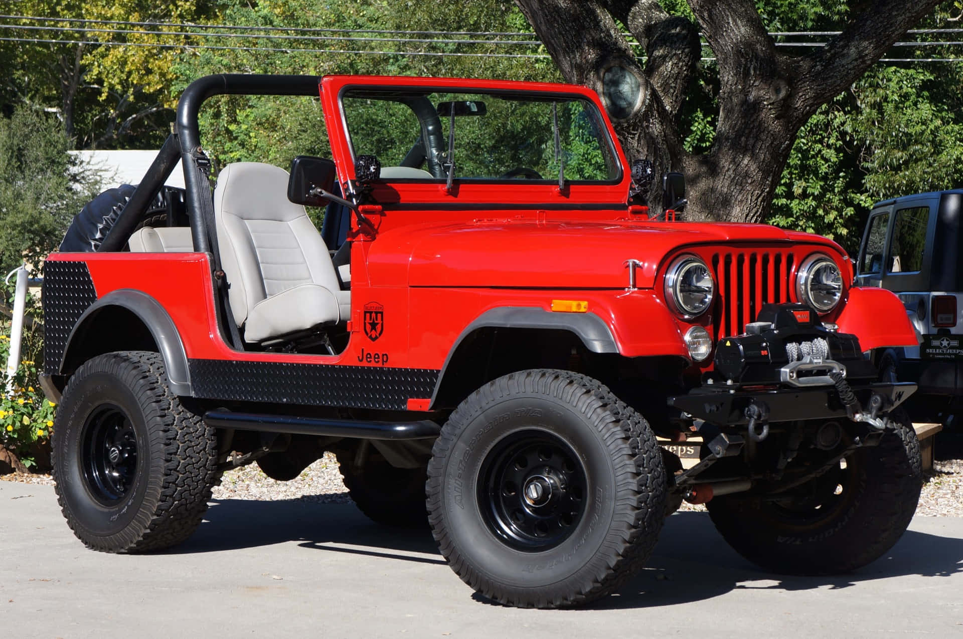 Jeep Cj 桌布 壁紙 — 一輛鮮紅色的經典Jeep Wrangler，帶有黑色飾邊和大尺寸越野輪胎，在陽光明媚的日子裡停放在枝葉茂密的樹下。
