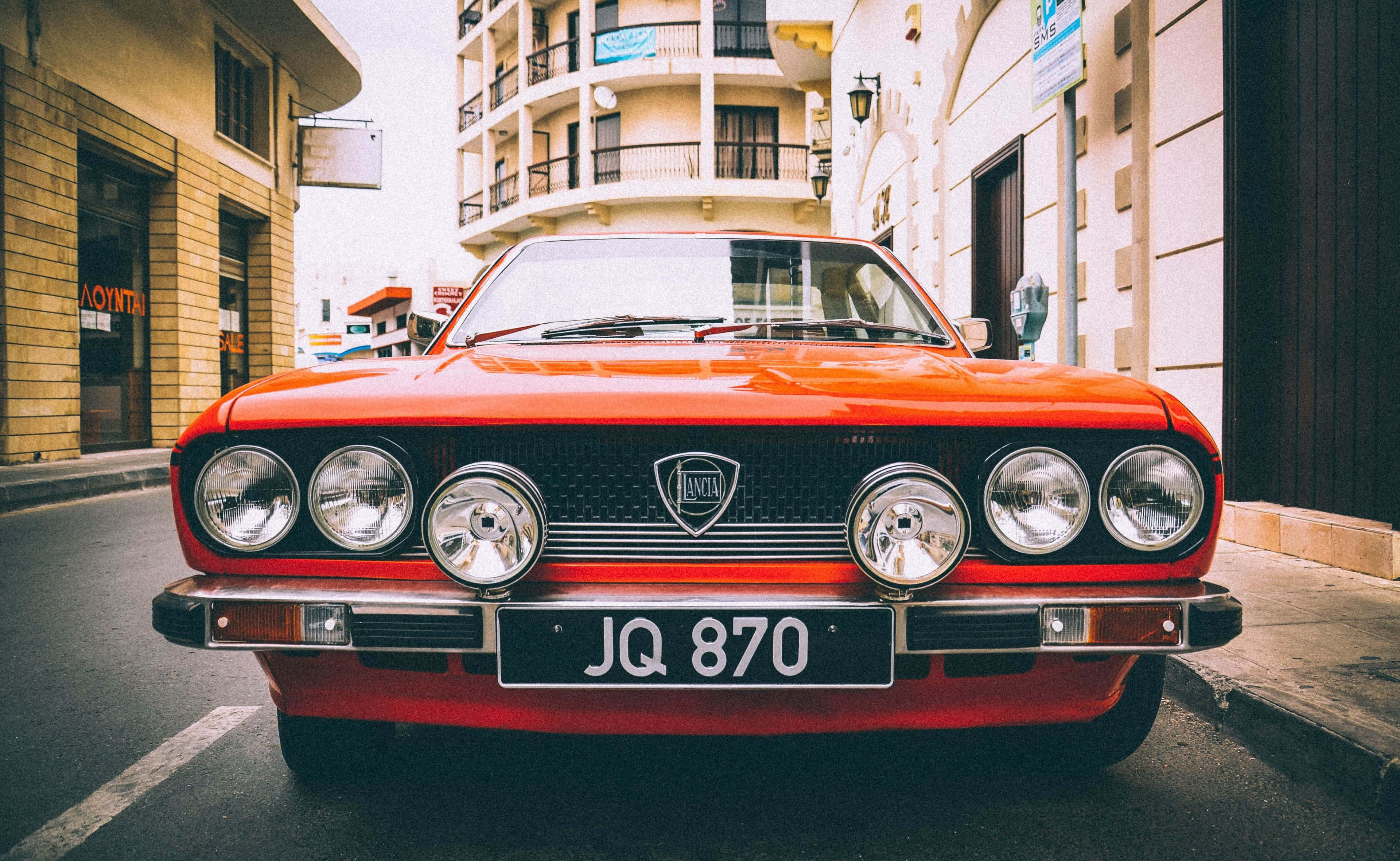 Lancia 桌布 壁紙 — 一輛鮮紅色的經典Lancia轎車，配備四盞頭燈，停放在陽光普照的城市街道上，兩旁是粉彩建築。