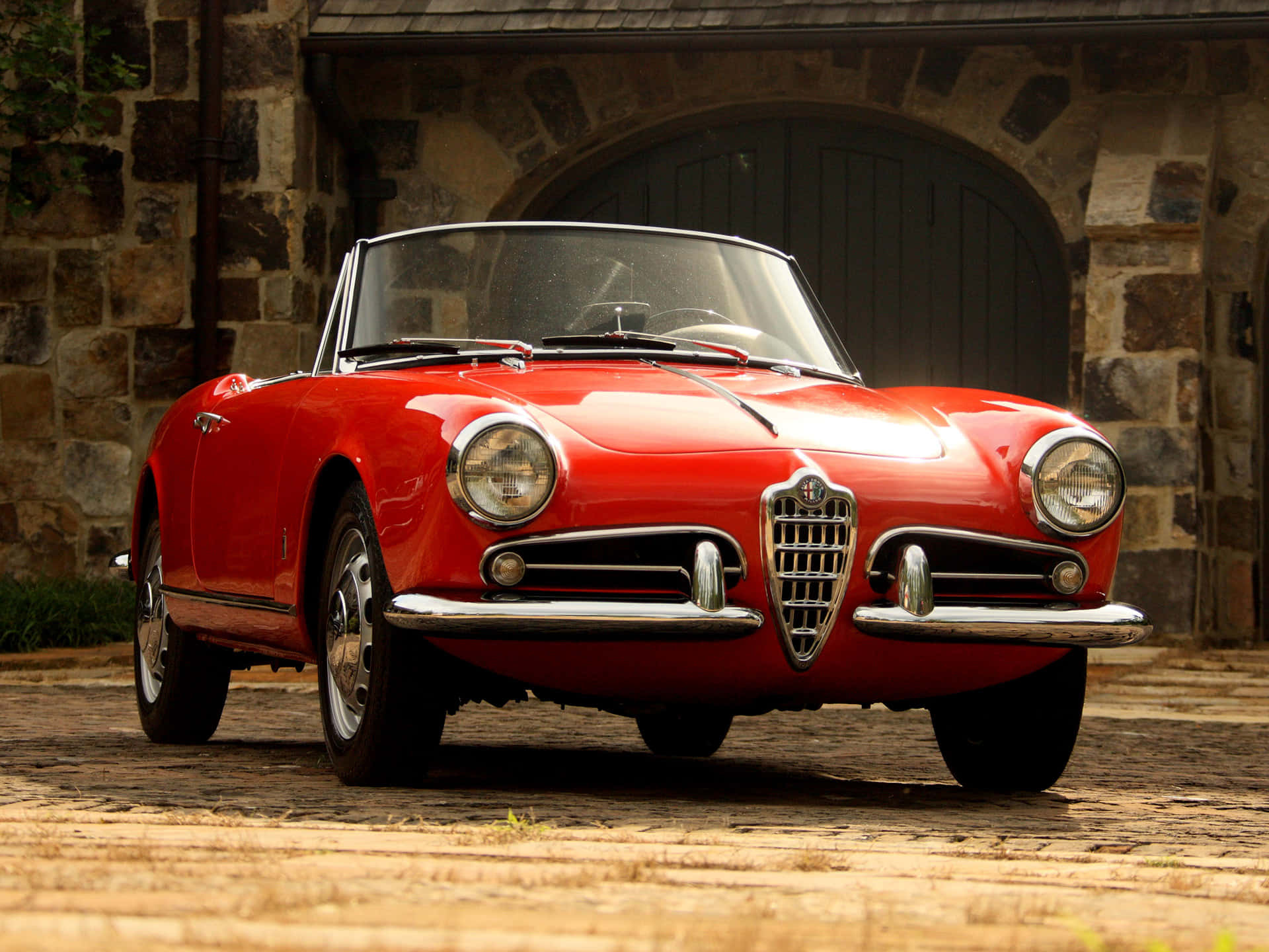 Alfa Romeo Spider 桌布 壁紙 — 一輛鮮紅色的經典愛快羅密歐Spider敞篷車停在石砌車庫前的鵝卵石上，車庫有拱形木門。