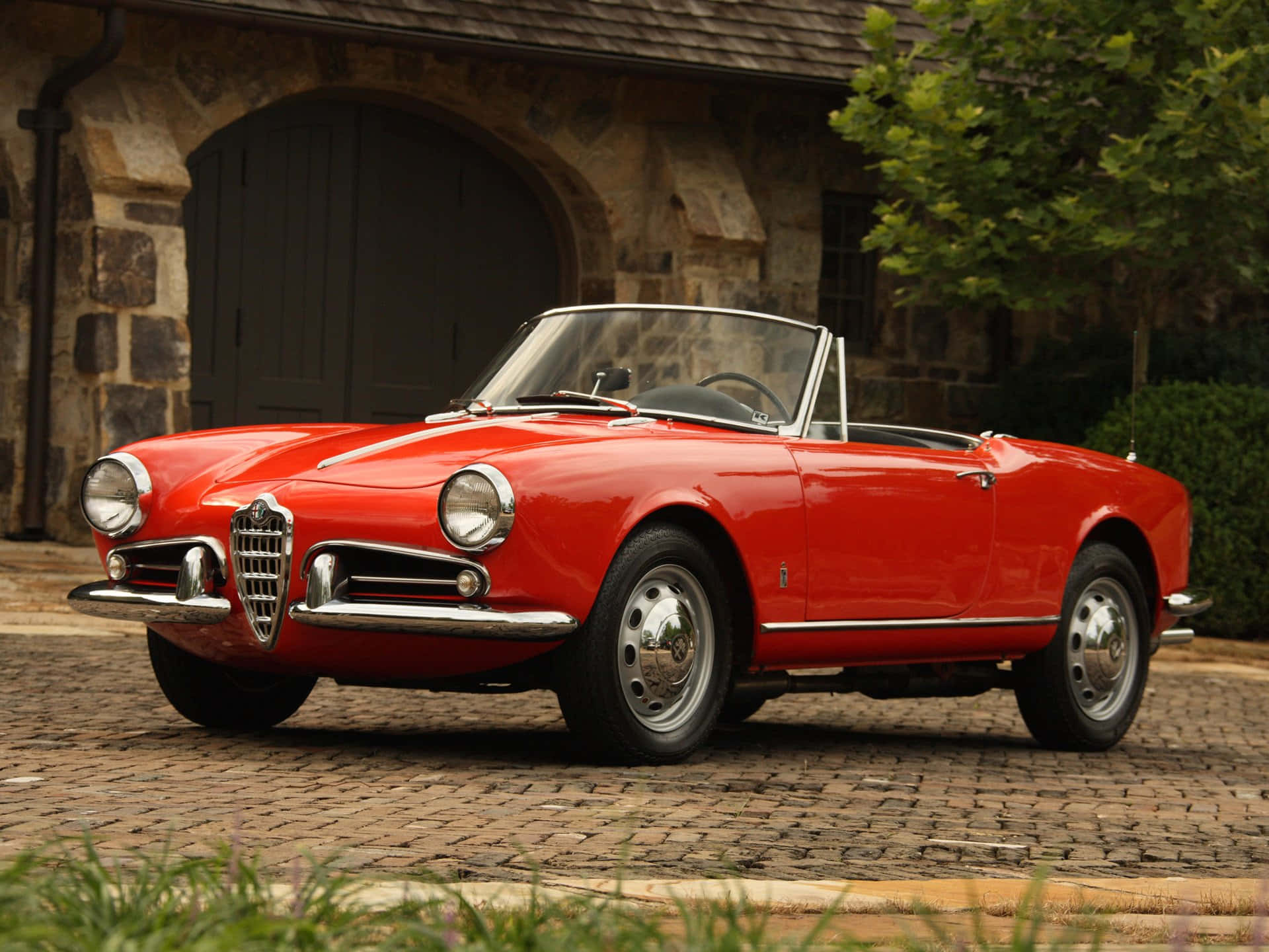 Alfa Romeo Spider 桌布 壁紙 — 一輛鮮紅色的經典愛快羅密歐Spider敞篷車停放在石砌建築前的鵝卵石車道上，周圍有綠植。