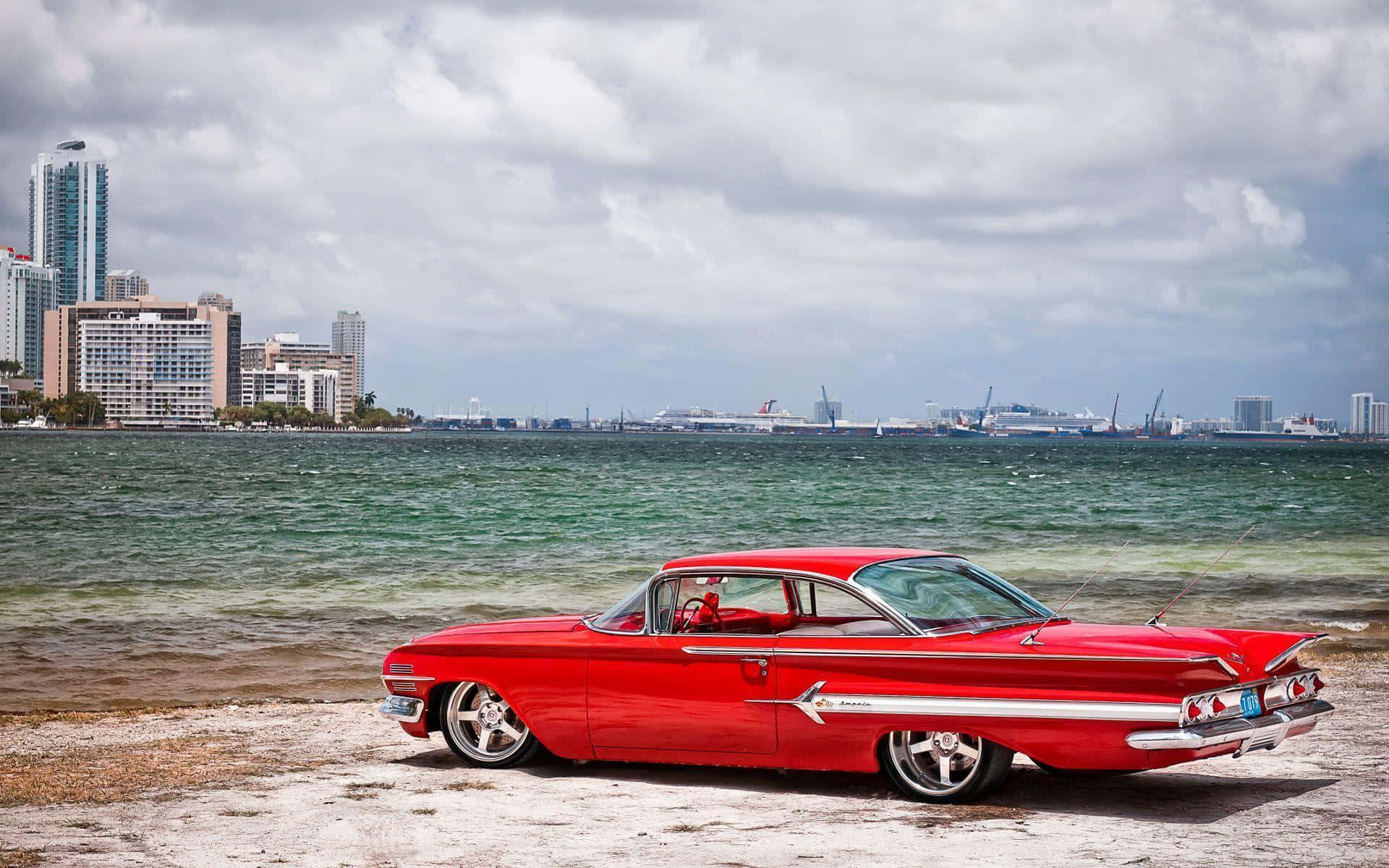 chevrolet impala 1967 桌布 壁紙 — 一輛鮮紅色的經典汽車停泊在沙灘上，背景是城市天際線和海洋，天空多雲。