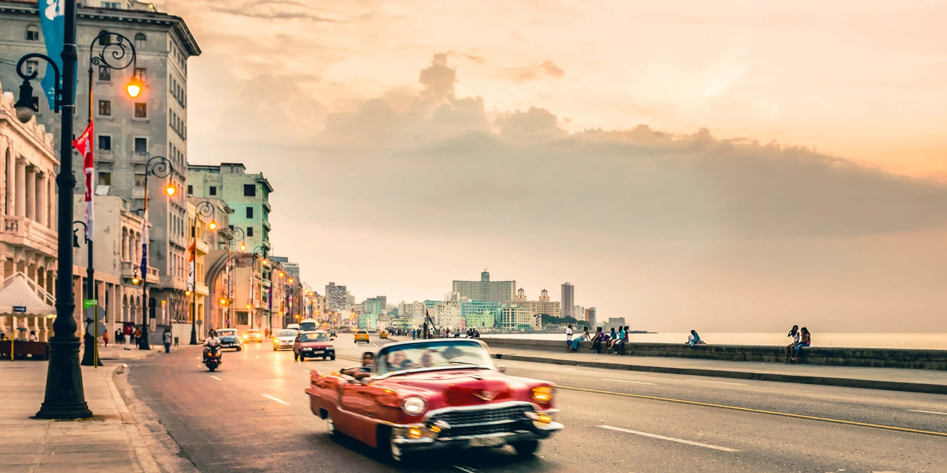 Cuba 桌布 壁紙 — 一輛鮮紅色的經典汽車在日落時分沿著哈瓦那的海濱大道行駛，兩旁是歷史建築和閃爍的路燈。
