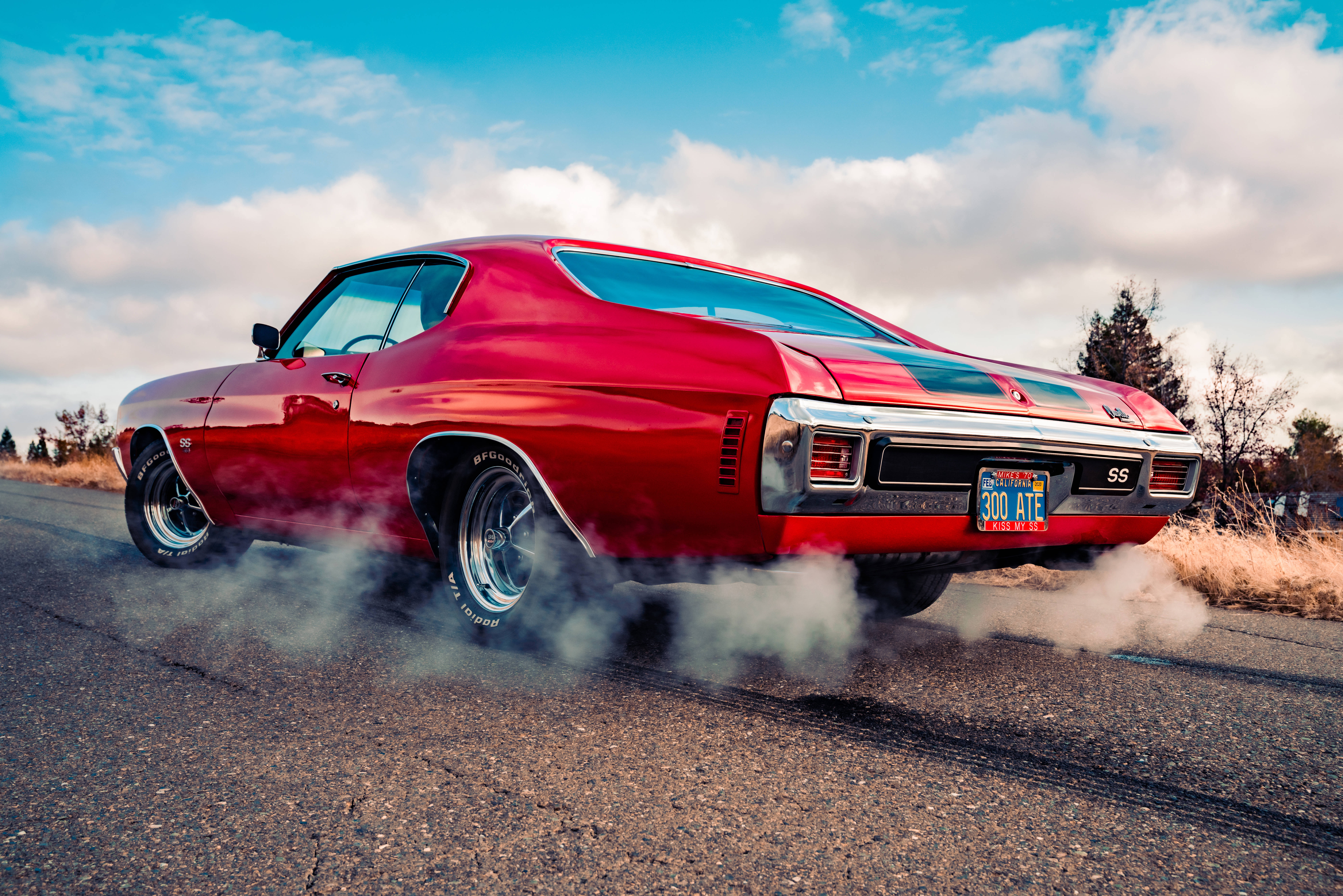 Chevrolet 桌布 壁紙 — 一輛鮮紅色的經典肌肉車在破裂的柏油路上燒胎，煙霧瀰漫，背景是晴朗藍天和 fluffy 雲朵。