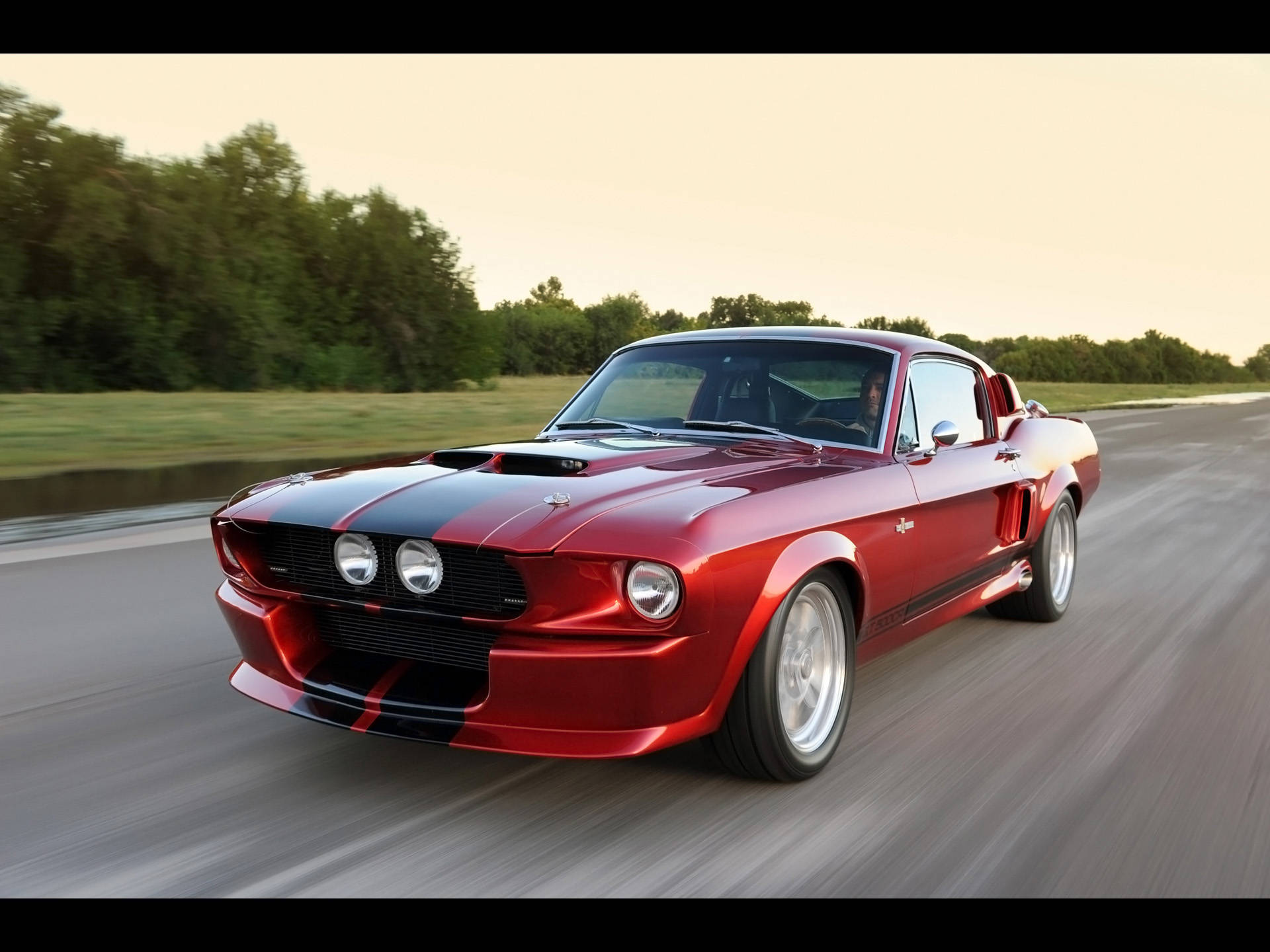 shelby iPhone 桌布 壁紙 — 一輛鮮紅色的經典肌肉車帶有黑色賽車條紋，在日落時分沿著鄉村道路飛馳。