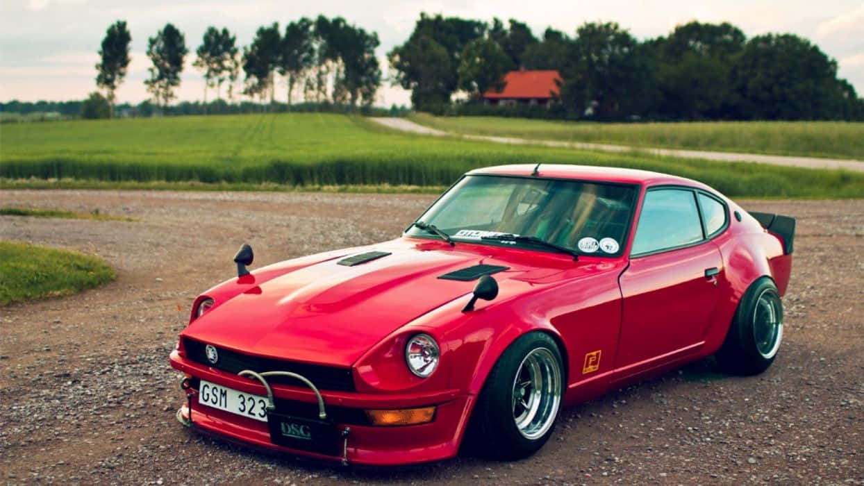 Datsun 桌布 壁紙 — 一輛鮮紅色的經典跑車，配備寬輪，停泊在碎石路上，旁邊是茂密的綠色田野，天空多雲。