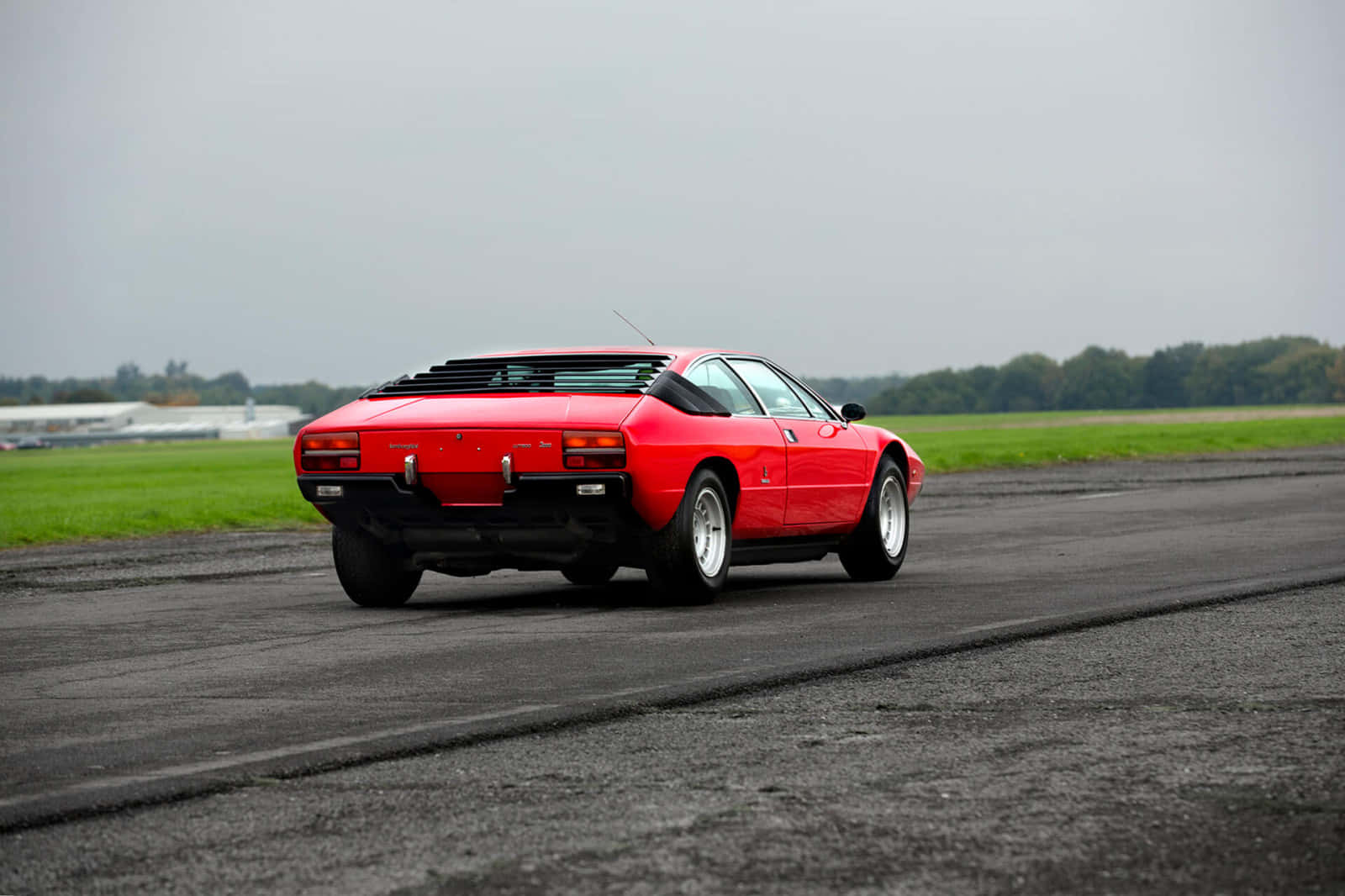 Lamborghini Urraco 桌布 壁紙 — 一輛鮮紅色的經典跑車在陰天之下，沿著濕漉漉的瀝青跑道行駛，兩旁是翠綠的田野。