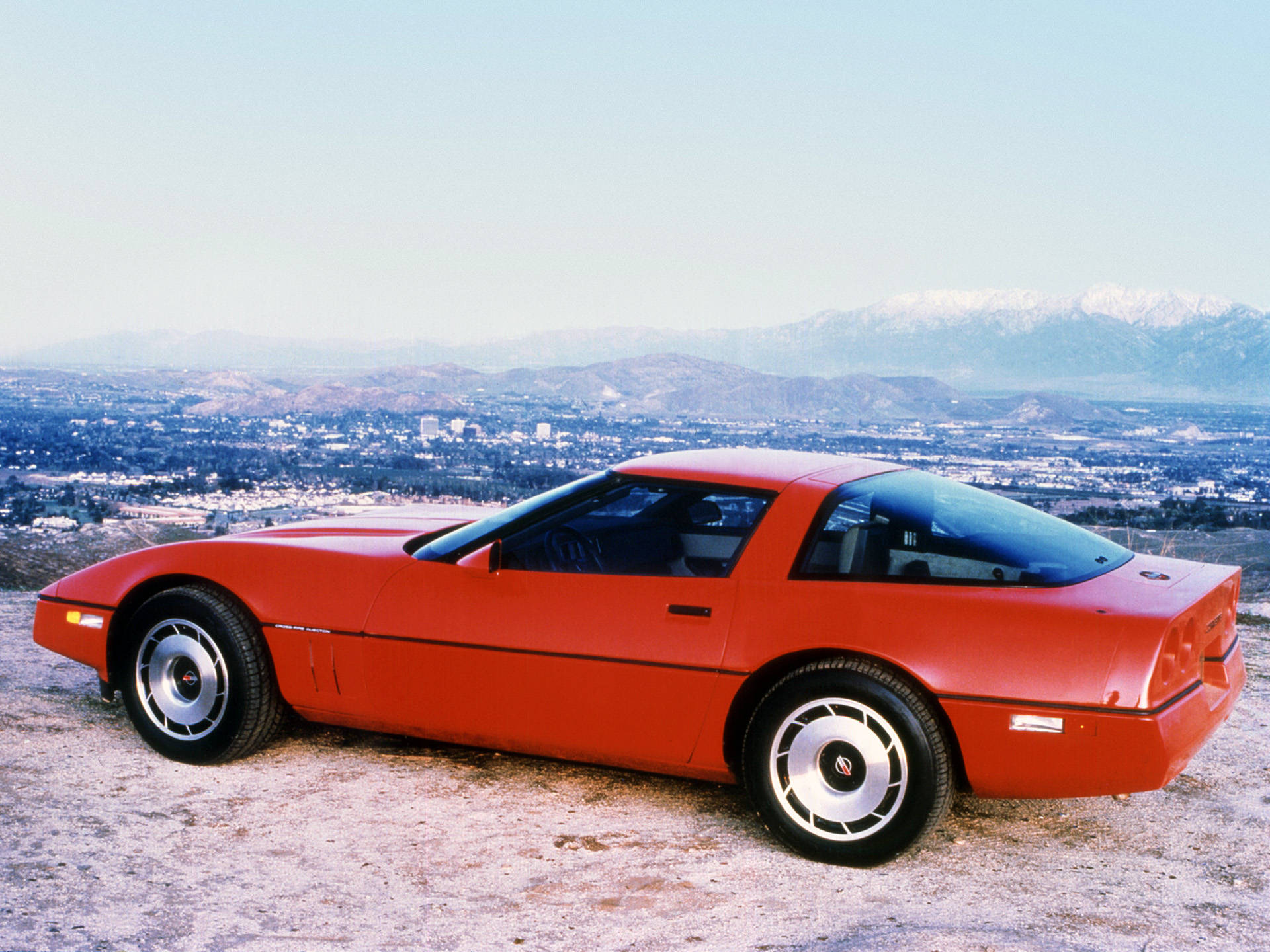 corvette 桌布 壁紙 — 一輛鮮紅色的經典跑車棲息在山丘上，俯瞰著廣闊的城市，遠處是白雪覆蓋的山脈。