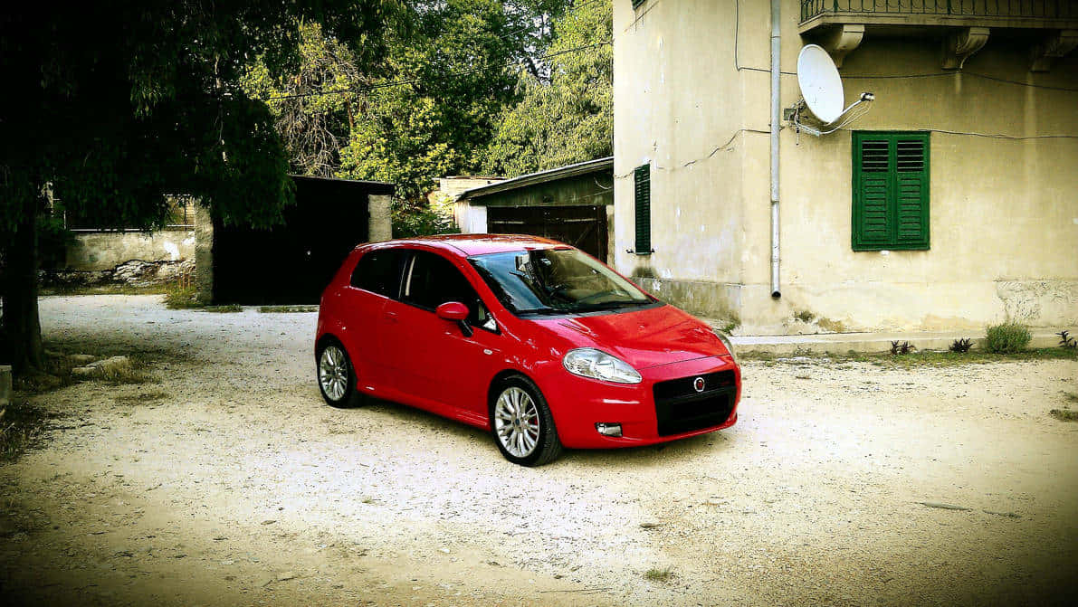 Fiat Punto 桌布 壁紙 — 一輛鮮紅色的緊湊型汽車停放在塵土飛揚的碎石車道上，旁邊是一座帶有綠色百葉窗的風化米色建築。