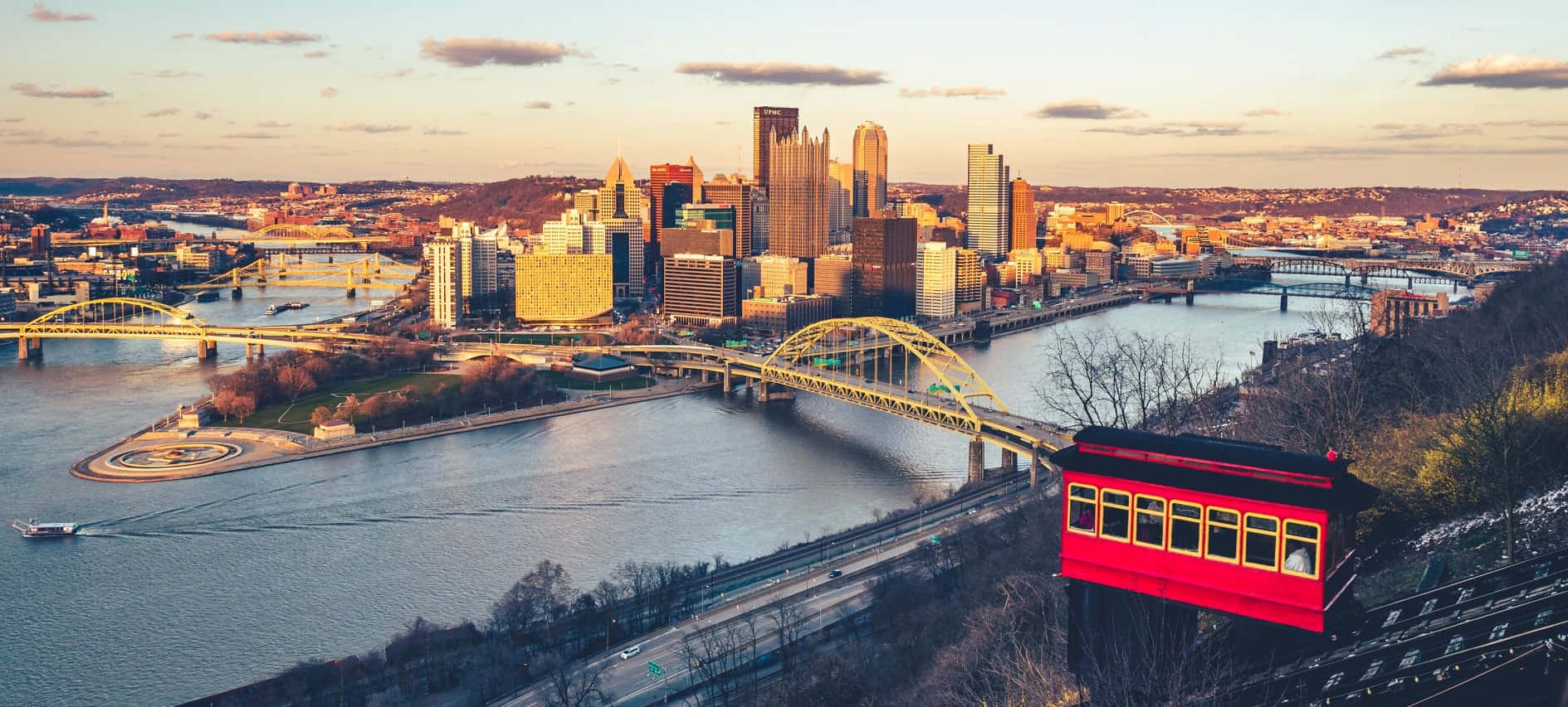 Pittsburgh Skyline 桌布 壁紙 — 一輛鮮紅色的纜車俯瞰著廣闊的城市景觀，正值黃金時刻，河流、橋樑和摩天大樓沐浴在溫暖的陽光中。