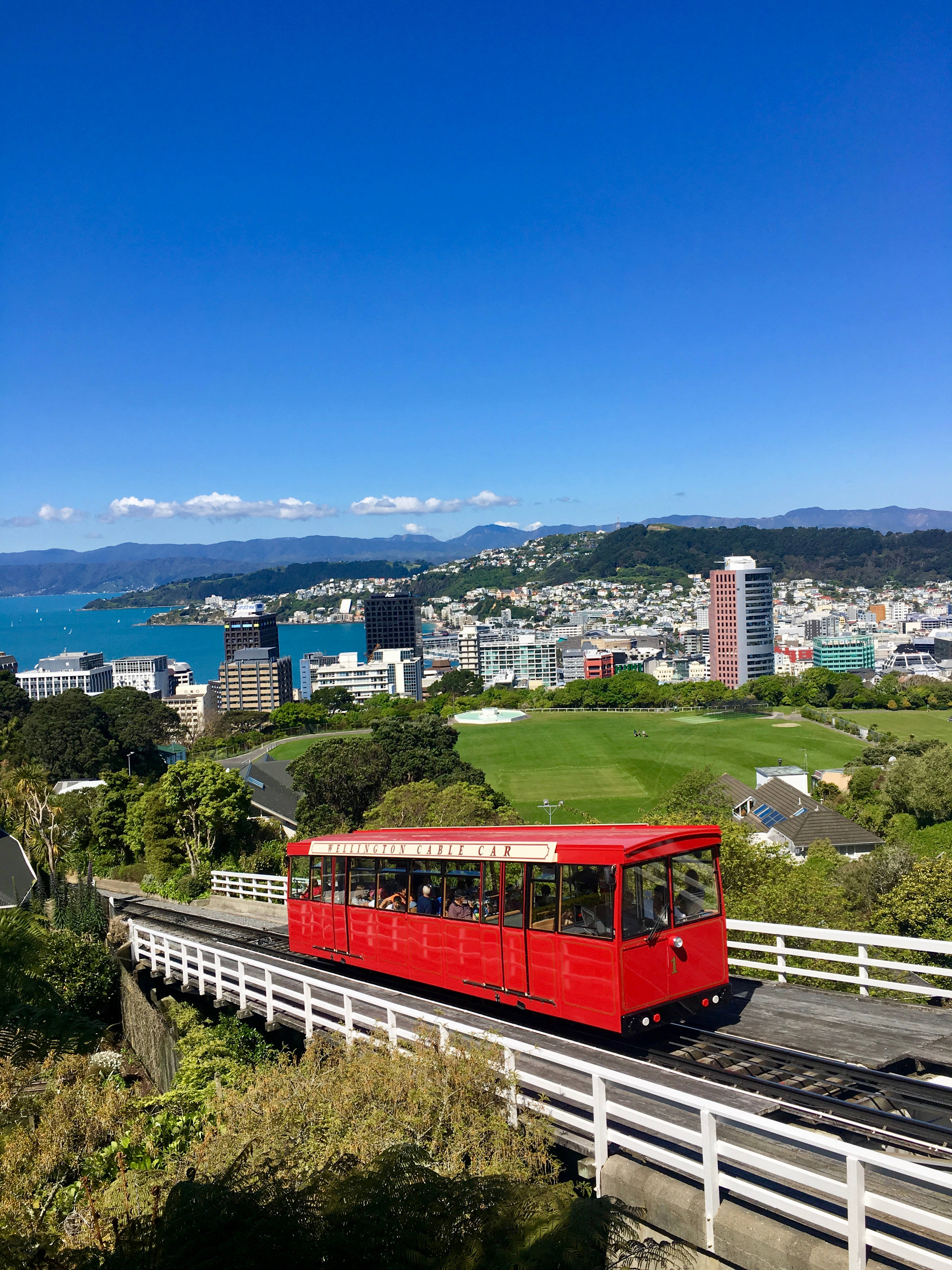 火車 桌布 壁紙 — 一輛鮮紅色的纜車沿著木製軌道滑行，俯瞰著一個充滿活力的沿海城市，擁有碧綠的海水和遠處的山脈，在晴朗的藍天下。