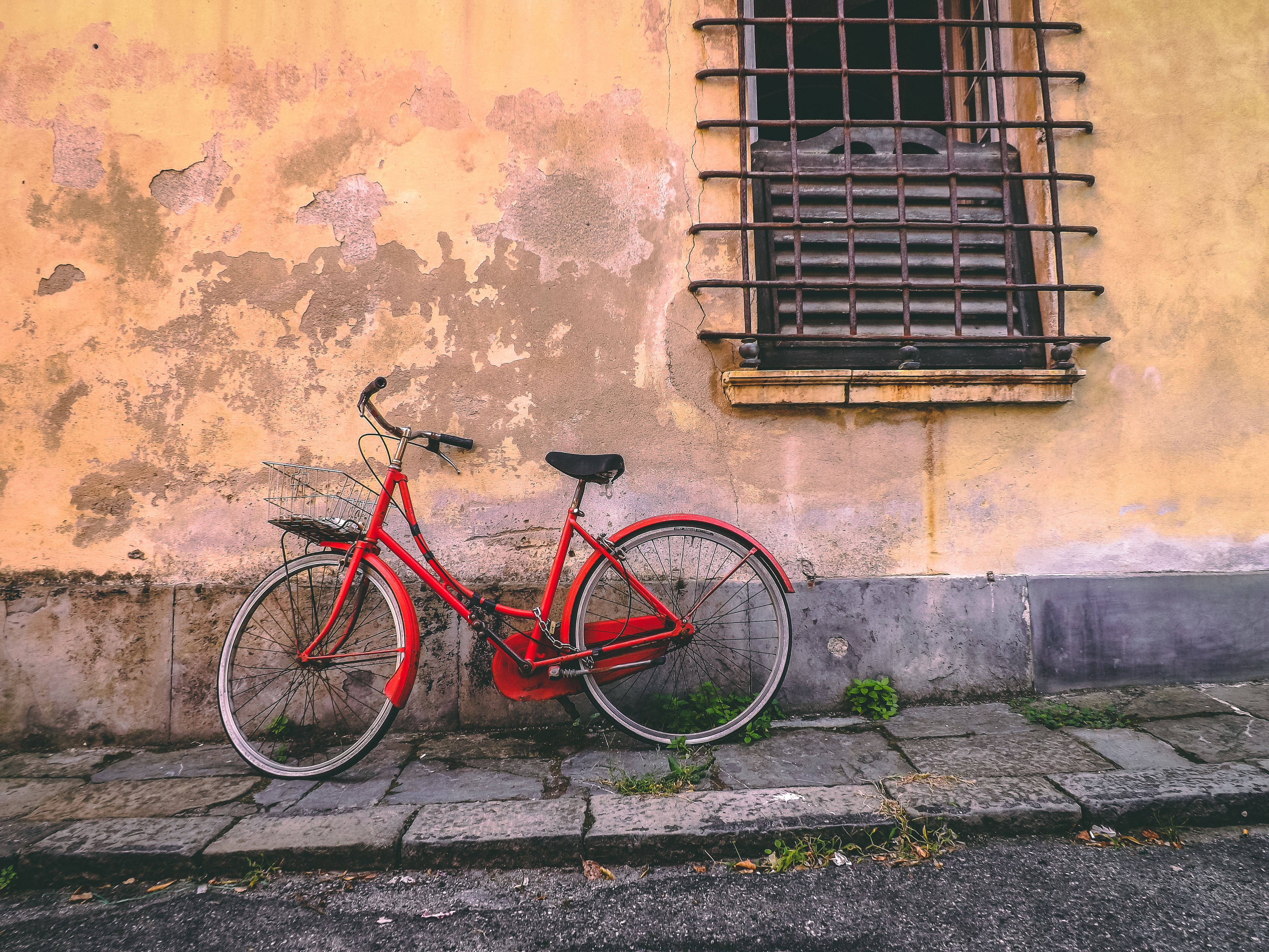 Bicycle 桌布 壁紙 — 一輛鮮紅色的腳踏車靠在風化的赭色牆上，旁邊是帶有鐵欄的窗戶，位於寧靜的城市人行道上。