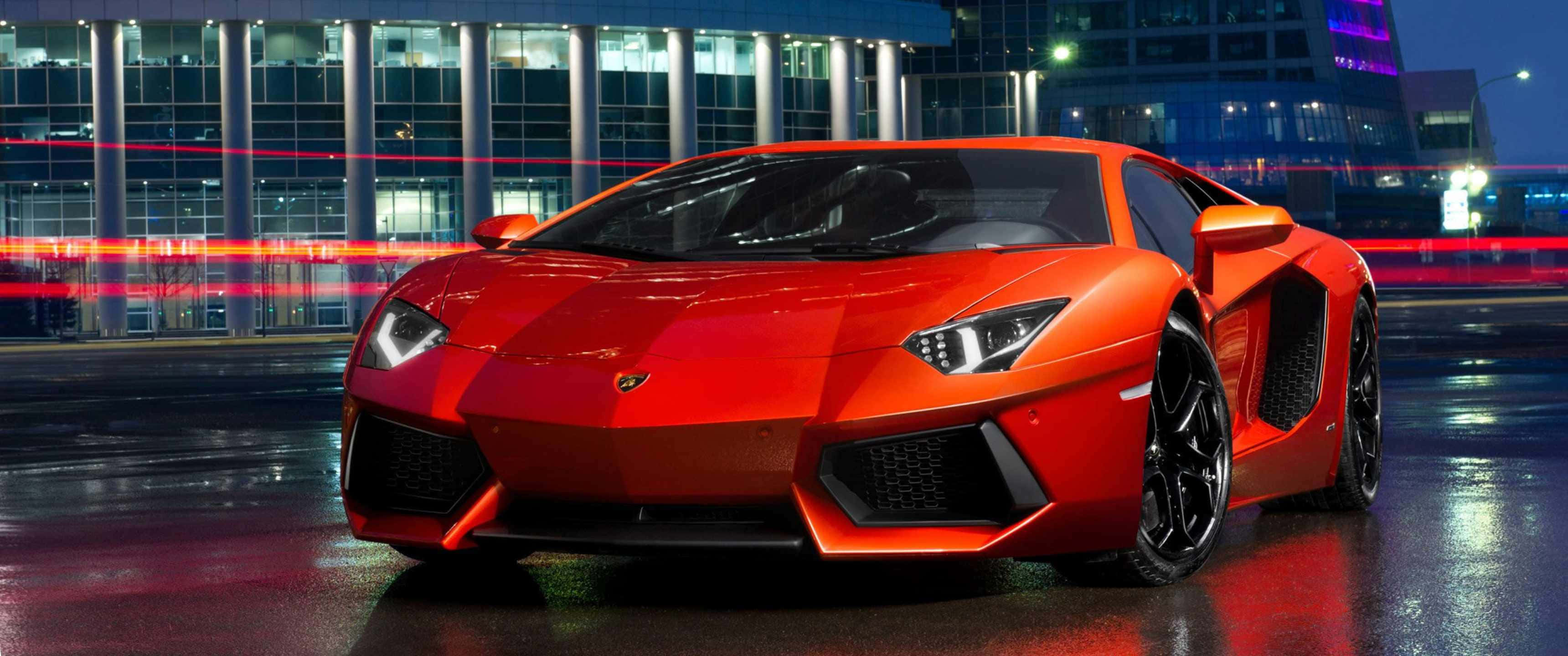 3440x1440p Lamborghini 桌布 壁紙 — 一輛鮮紅色的藍寶堅尼Aventador在潮濕的夜街上，於城市燈光下閃耀，背後有模糊的車流線條。