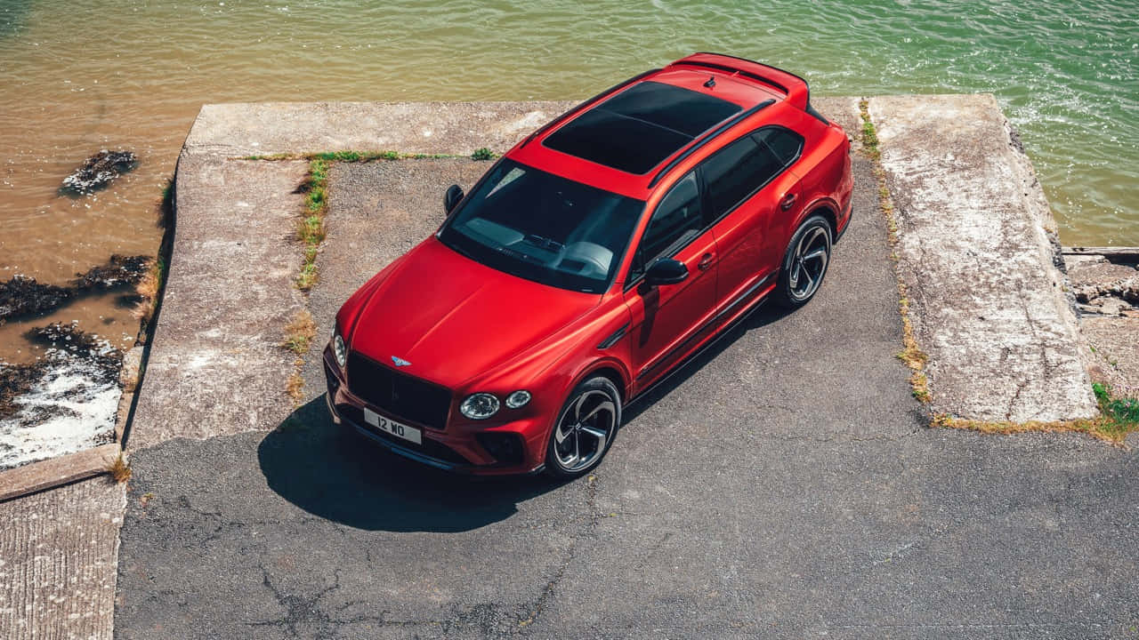 Bentley Bentayga 桌布 壁紙 — 一輛鮮紅色的賓利 Bentayga SUV 停泊在風化的混凝土碼頭上，旁邊是波光粼粼的水面，在明亮的日光下。