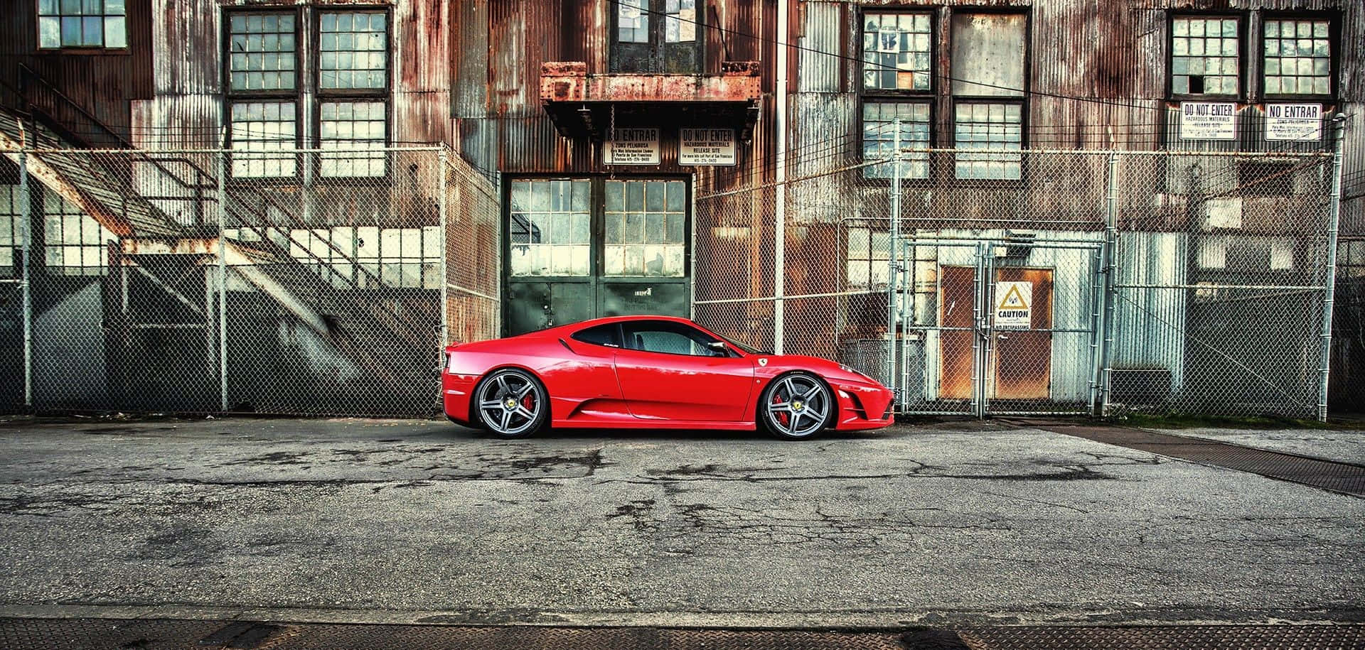 Ferrari 360 Modena 桌布 壁紙 — 一輛鮮紅色的跑車停放在風化的工業建築前，周圍有生鏽的金屬和鐵絲網圍欄。