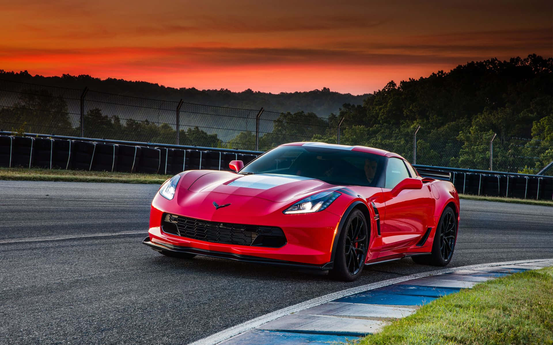 Chevrolet Corvette Grand Sport 桌布 壁紙 — 一輛鮮紅色的跑車在日落時分沿著賽道飛馳，背景是火紅的橙色天空和綠色山丘。