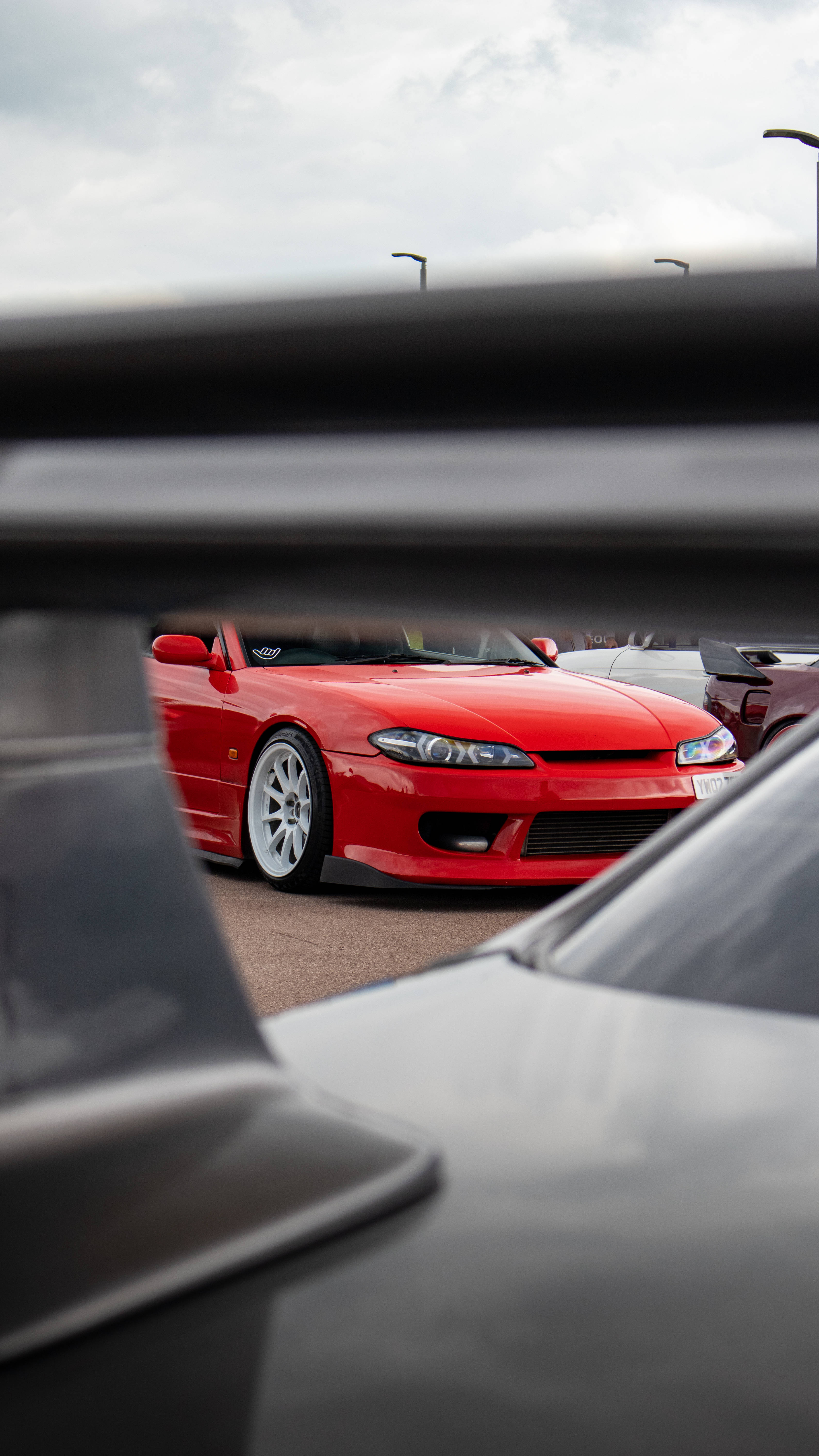 Rx7 iPhone 桌布 壁紙 — 一輛鮮紅色的跑車在陰天之下閃閃發光，由停車場中另一輛車的模糊引擎蓋框住。