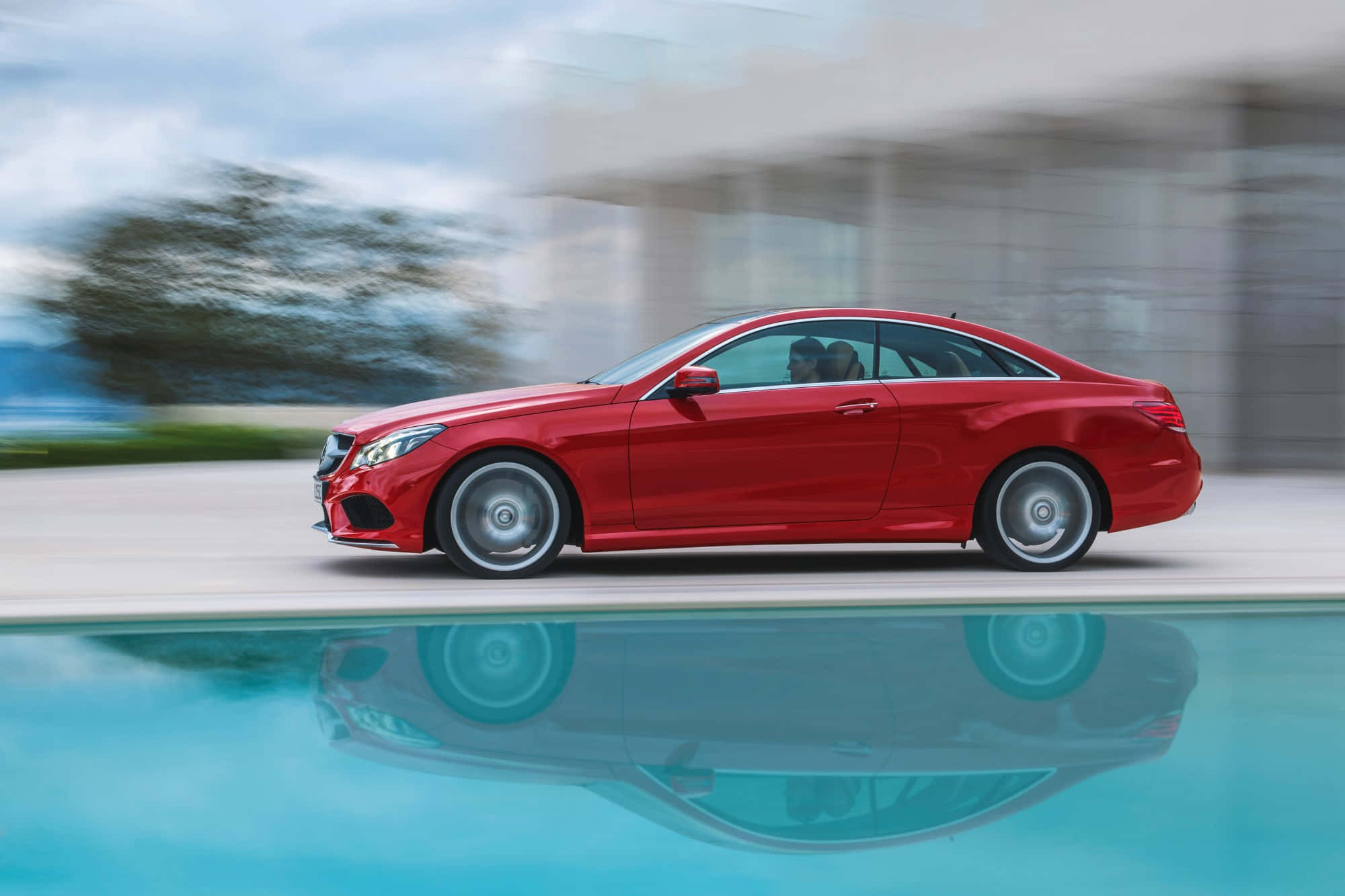 Mercedes Benz Clase E 桌布 壁紙 — 一輛鮮紅色的跑車高速駛過泳池，其倒影在模糊的現代背景中於水面上閃爍。