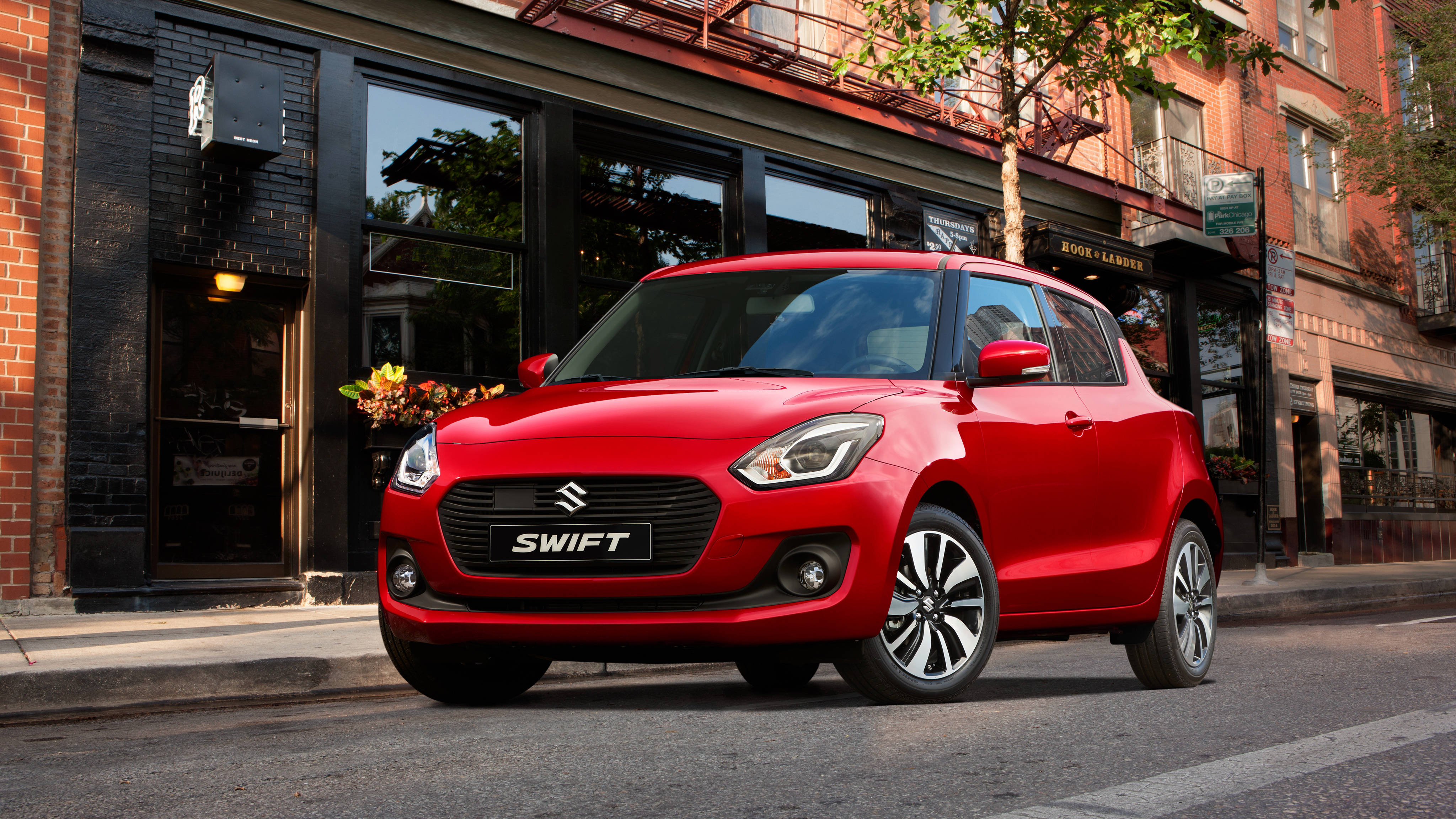 Suzuki 桌布 壁紙 — 一輛鮮紅色的鈴木 Swift 停泊在城市的街道上，旁邊是帶有商店門面和秋葉的磚砌建築。