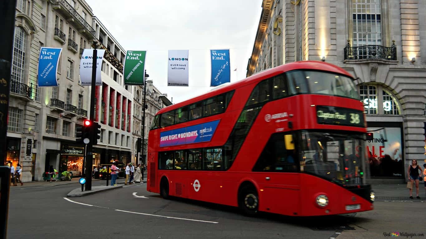 Bus 桌布 壁紙 — 一輛鮮紅色的雙層巴士在倫敦一條繁忙的街道上轉彎，街道兩旁是歷史建築和西區的橫幅。