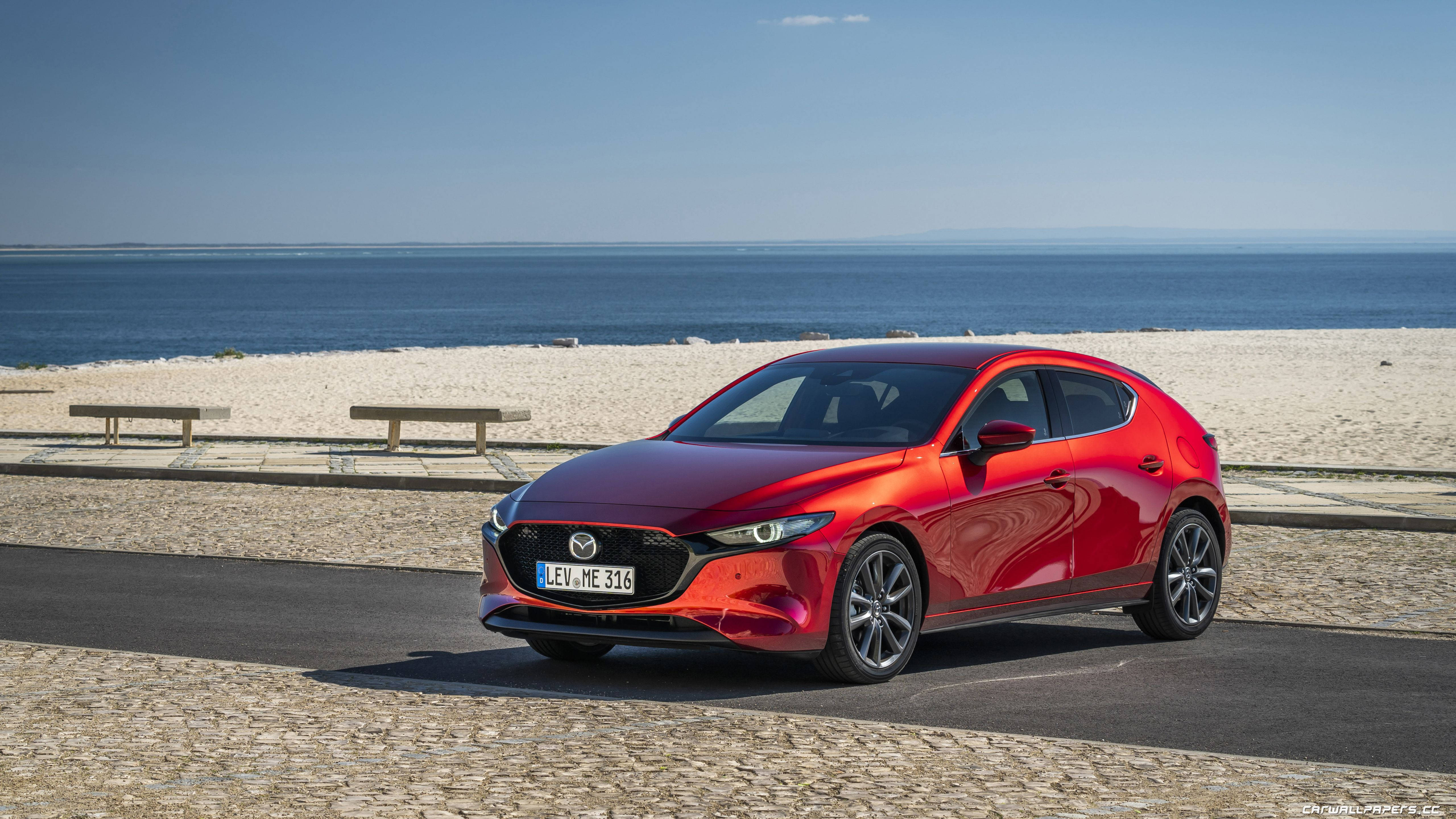 Mazda 桌布 壁紙 — 一輛鮮紅色的馬自達3掀背車在沿海道路上閃閃發光，旁邊是沙灘和寧靜的藍色大海，天空晴朗。