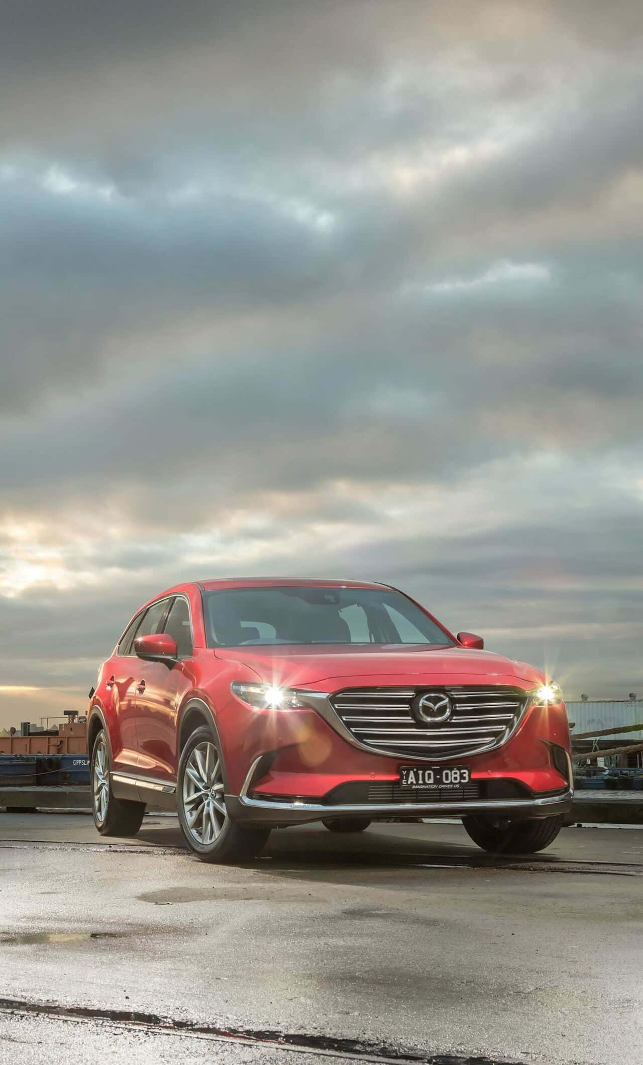 Mazda Cx-9 桌布 壁紙 — 一輛鮮紅色的馬自達CX-9 SUV在戲劇性的多雲天空下閃閃發光，車頭燈亮著，停在濕漉漉的工業碼頭上。