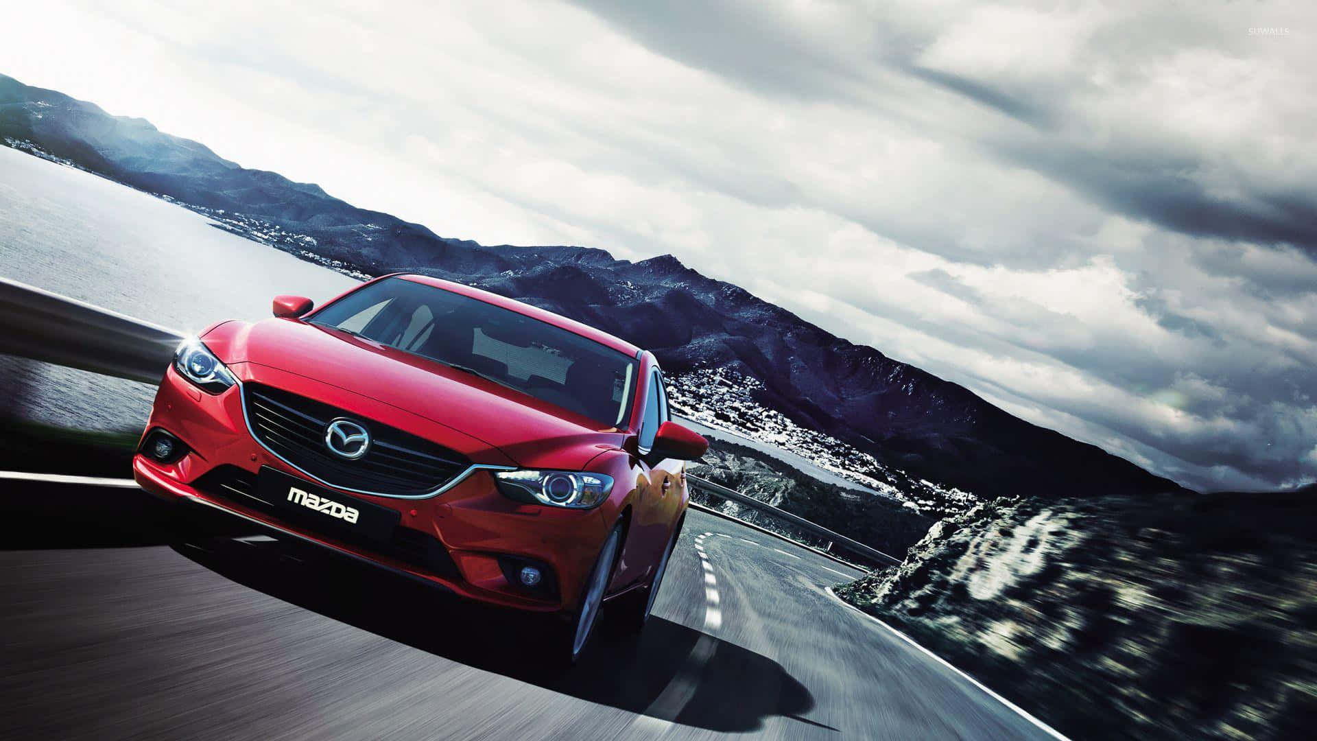 Mazda 6 桌布 壁紙 — 一輛鮮紅色的馬自達沿著蜿蜒的海岸道路疾馳，背景是山脈和多雲的天空。