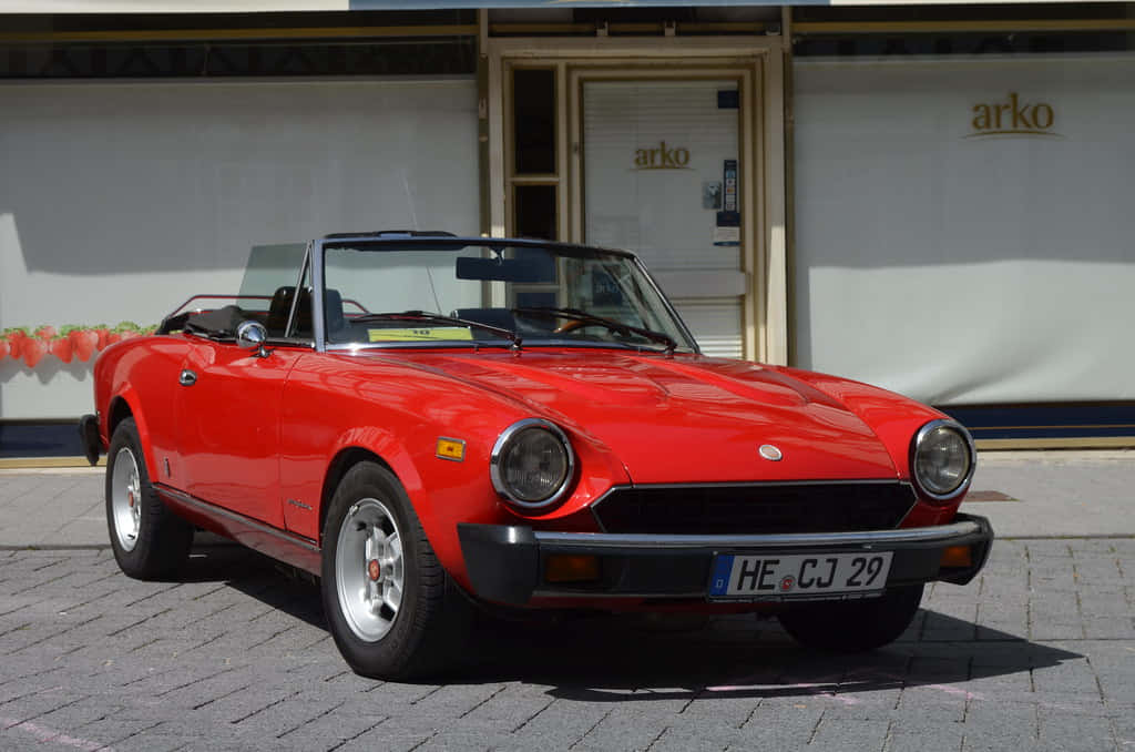 Fiat 124 Spider 桌布 壁紙 — 一輛鮮紅色經典敞篷跑車停放在陽光普照的鵝卵石街道上，前方是商店門面。