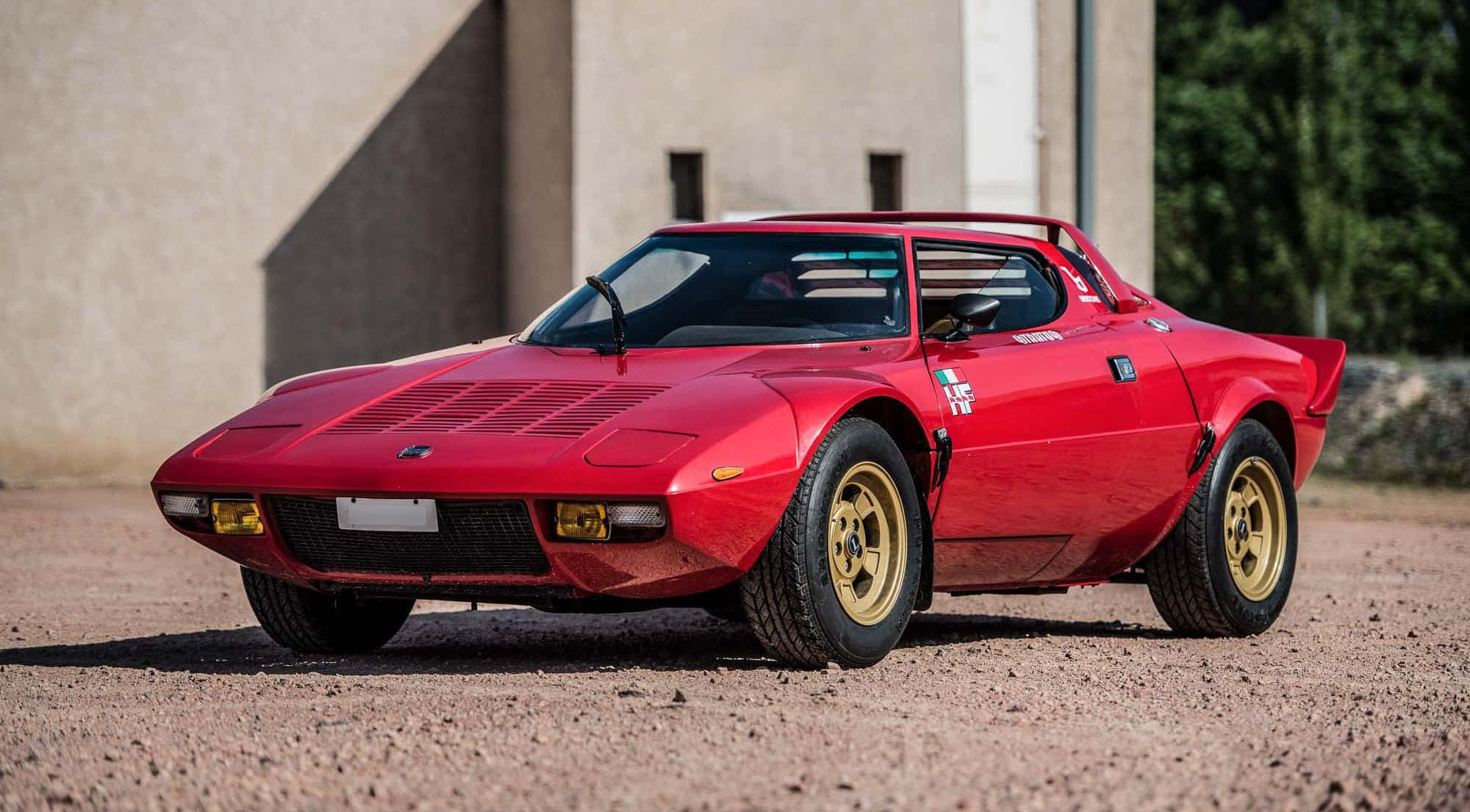 Lancia 桌布 壁紙 — 一輛鮮紅色經典跑車配金色輪圈，停泊在現代建築旁的碎石地上。