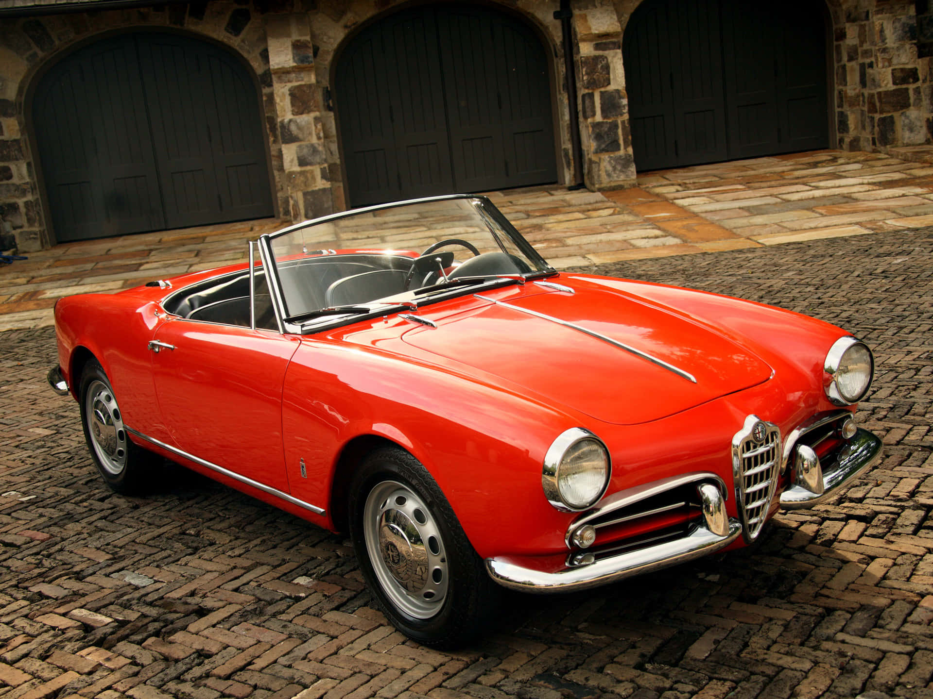 Alfa Romeo Spider 桌布 壁紙 — 一輛鮮紅色經典阿爾法羅密歐Spider敞篷車停放在石拱門前的鵝卵石庭院。
